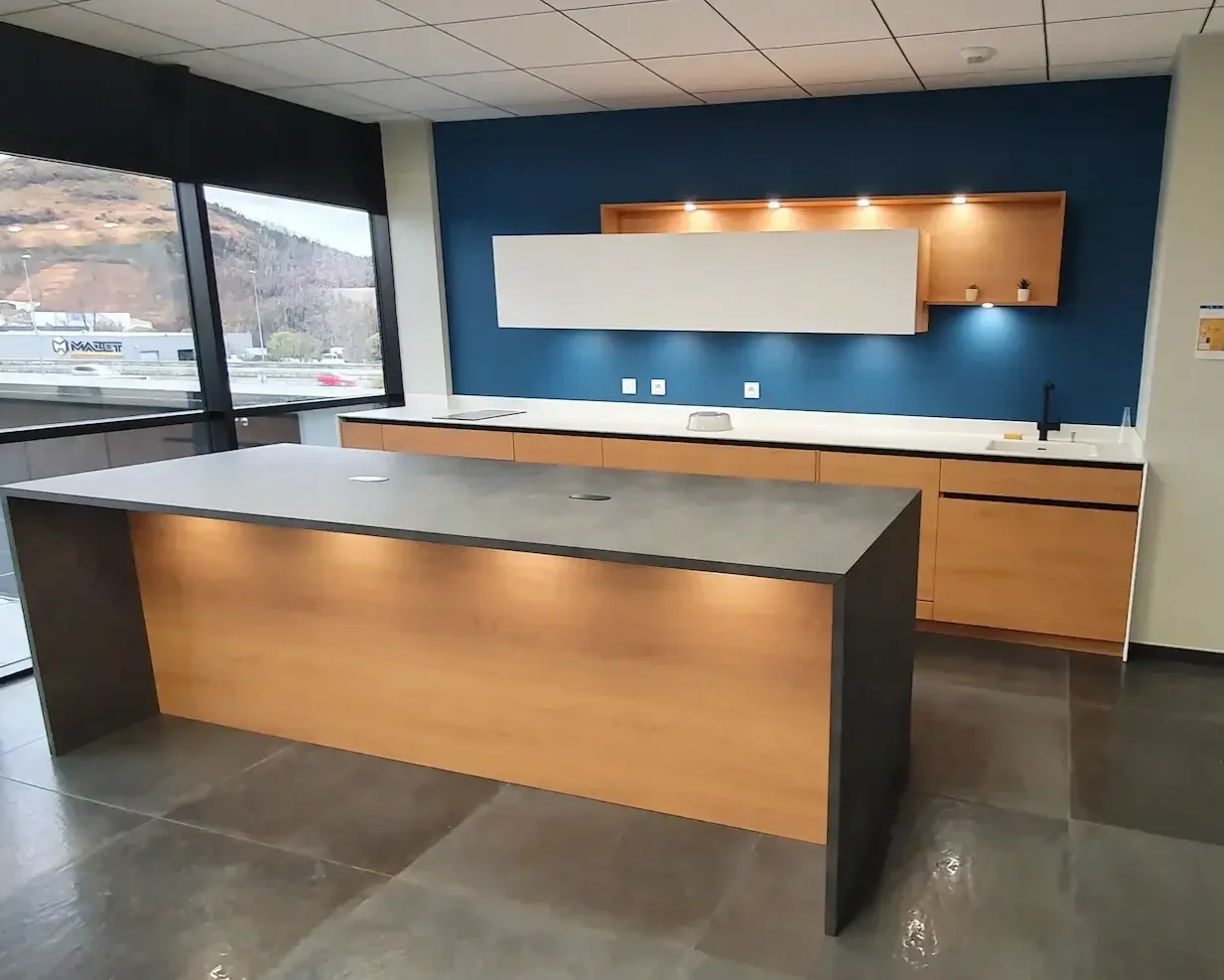 Cuisine moderne avec un plan de travail en pierre gris foncé, des armoires en bois clair, un mur bleu foncé, et une grande fenêtre donnant sur un paysage montagneux.