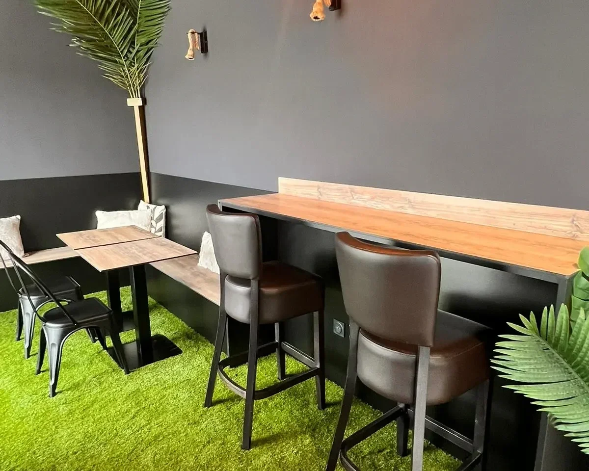 Espace de café avec des tables en bois, chaises en cuir marron, un mur gris foncé, plantes vertes, tapis vert.