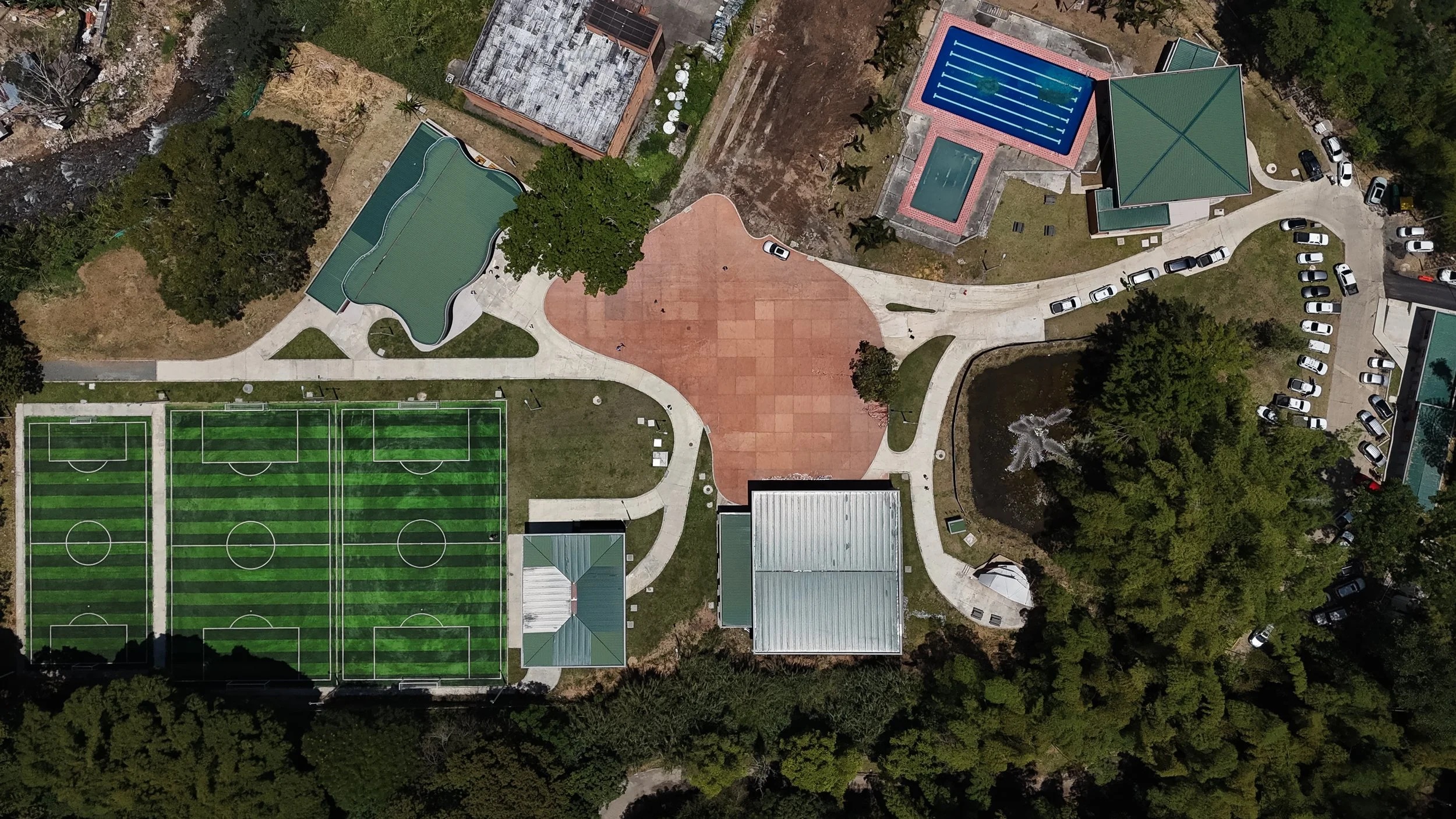 Vista aérea de un parque que incluye campos de fútbol, una piscina azul, un área de juegos con césped artificial, caminos pavimentados y áreas verdes con árboles, además de varios vehículos estacionados.