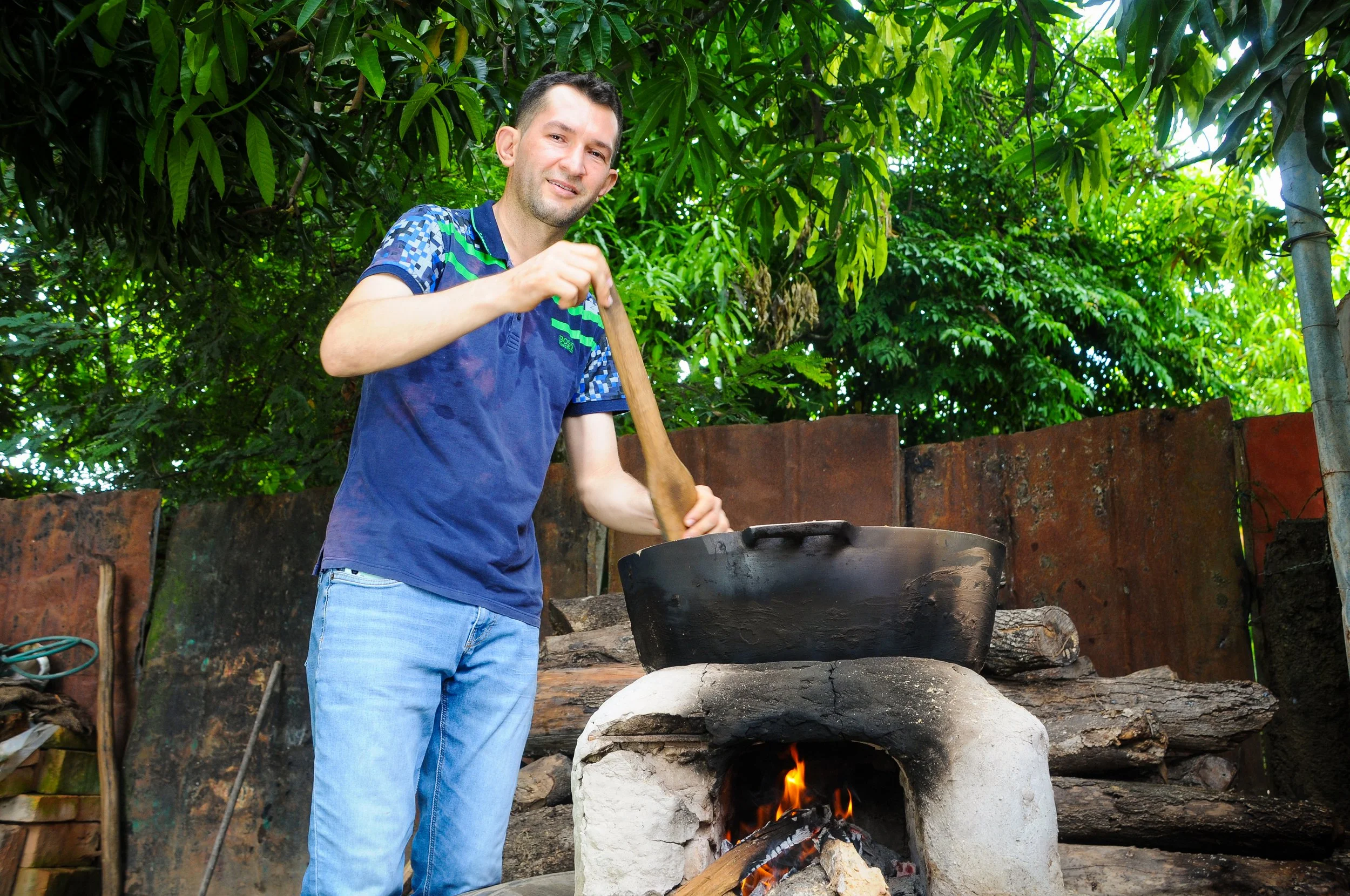 Hombre cocinando con un sartén grande sobre un horno de barro en un entorno exterior con vegetación densa.