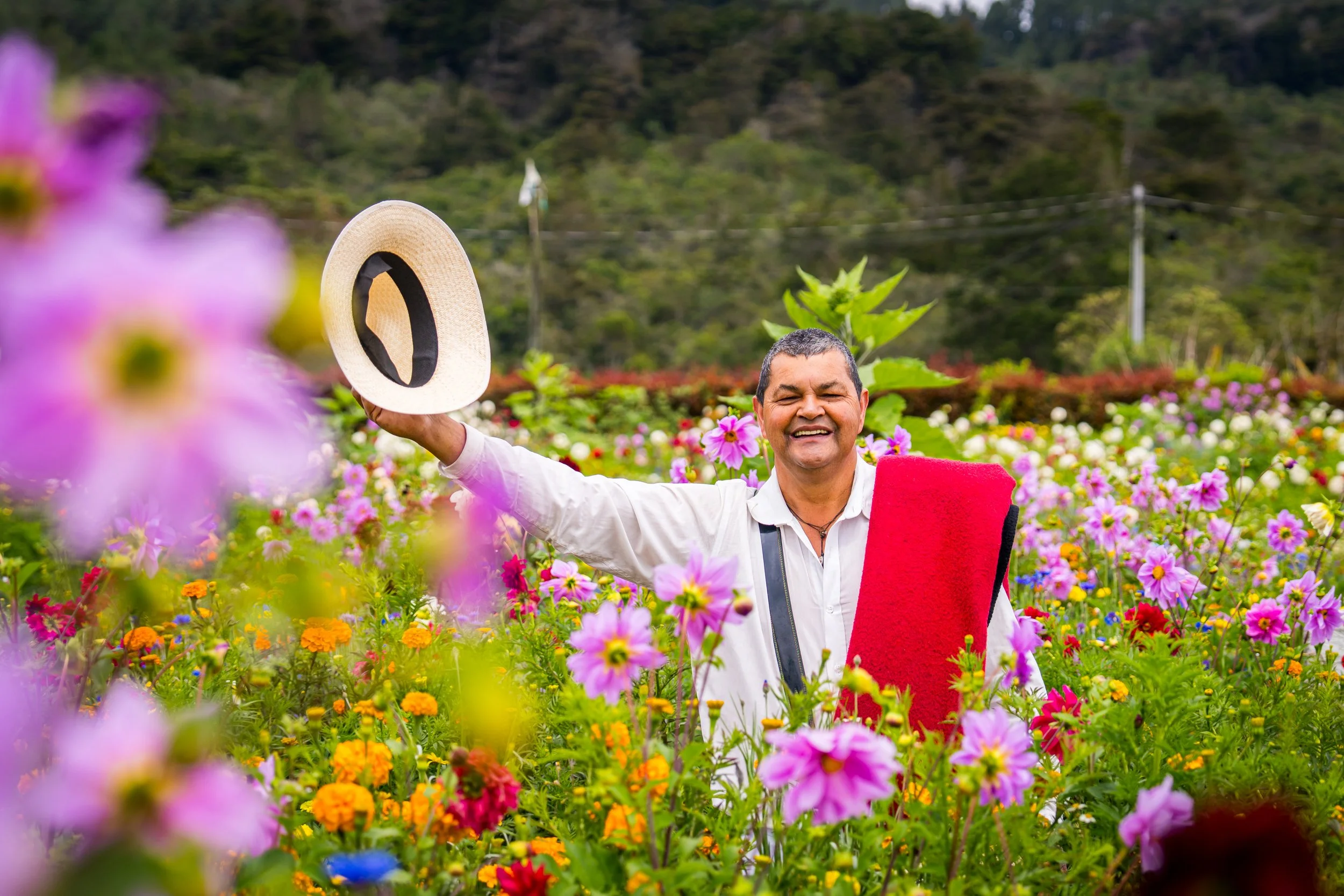 Hombre sonriendo en campo de flores de colores, llevando un sombrero y una manta roja sobre los hombros.