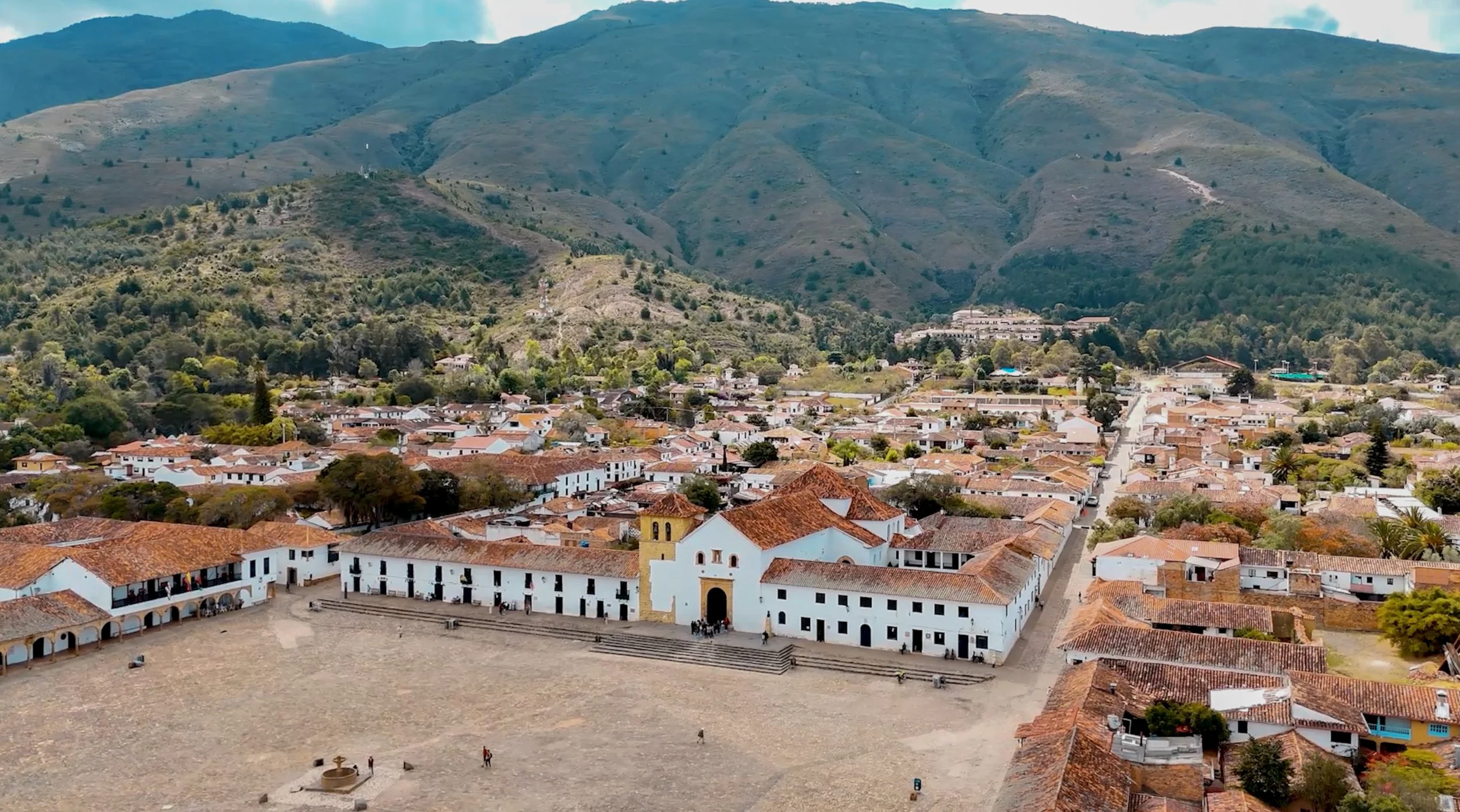 Vista panorámica de un pueblo colonial con calles empedradas, edificios de techos de teja y una plaza central, rodeada de montañas verdes.