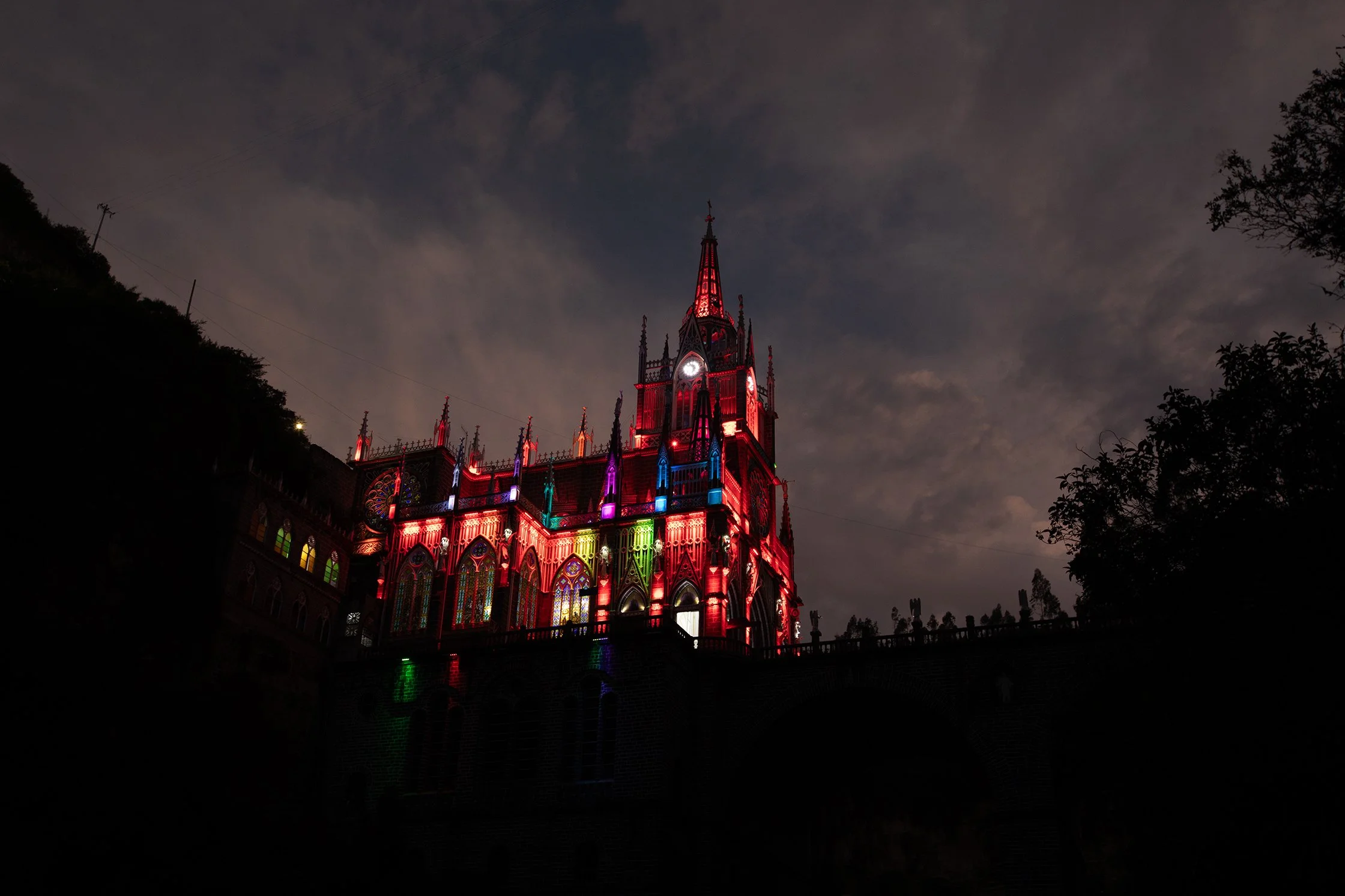 Una iglesia gótica iluminada con luces de colores en la noche, rodeada de árboles y un cielo nublado.