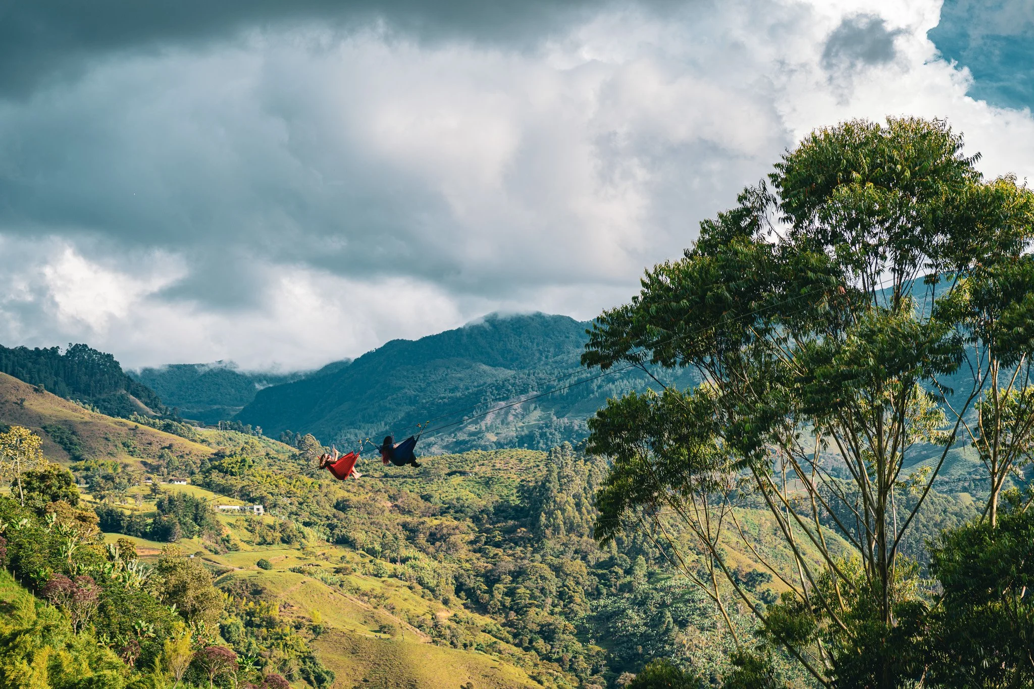 Paisaje montañoso con nubes grises, árboles verdes y colinas en lontananza, con dos hamacas colgadas en un cable entre árboles en primer plano.
