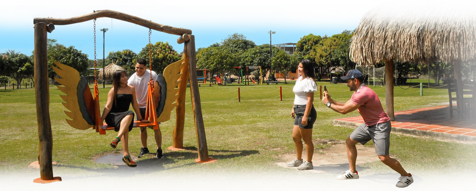Personas en un parque tomando fotos y jugando en un columpio con alas decorativas, en un día soleado.