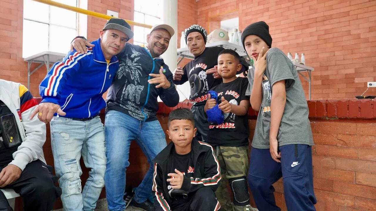 Grupo de seis personas, cuatro niños y dos adultos, posando en interior con paredes de ladrillo y ventanas grandes.