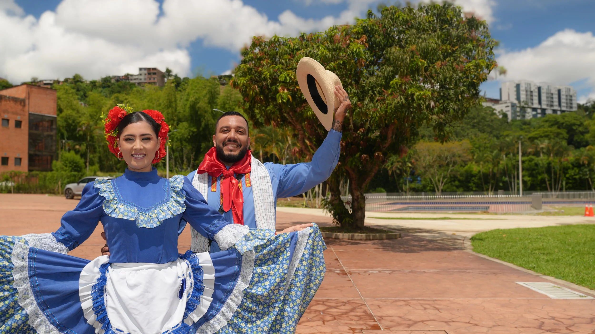 Pareja de bailarines con vestimenta tradicional colombiana en un parque con árboles y edificios en el fondo, ella lleva un vestido azul con volantes y flores en el cabello, él lleva camisa azul y una bufanda roja, y sostiene un sombrero en la mano.