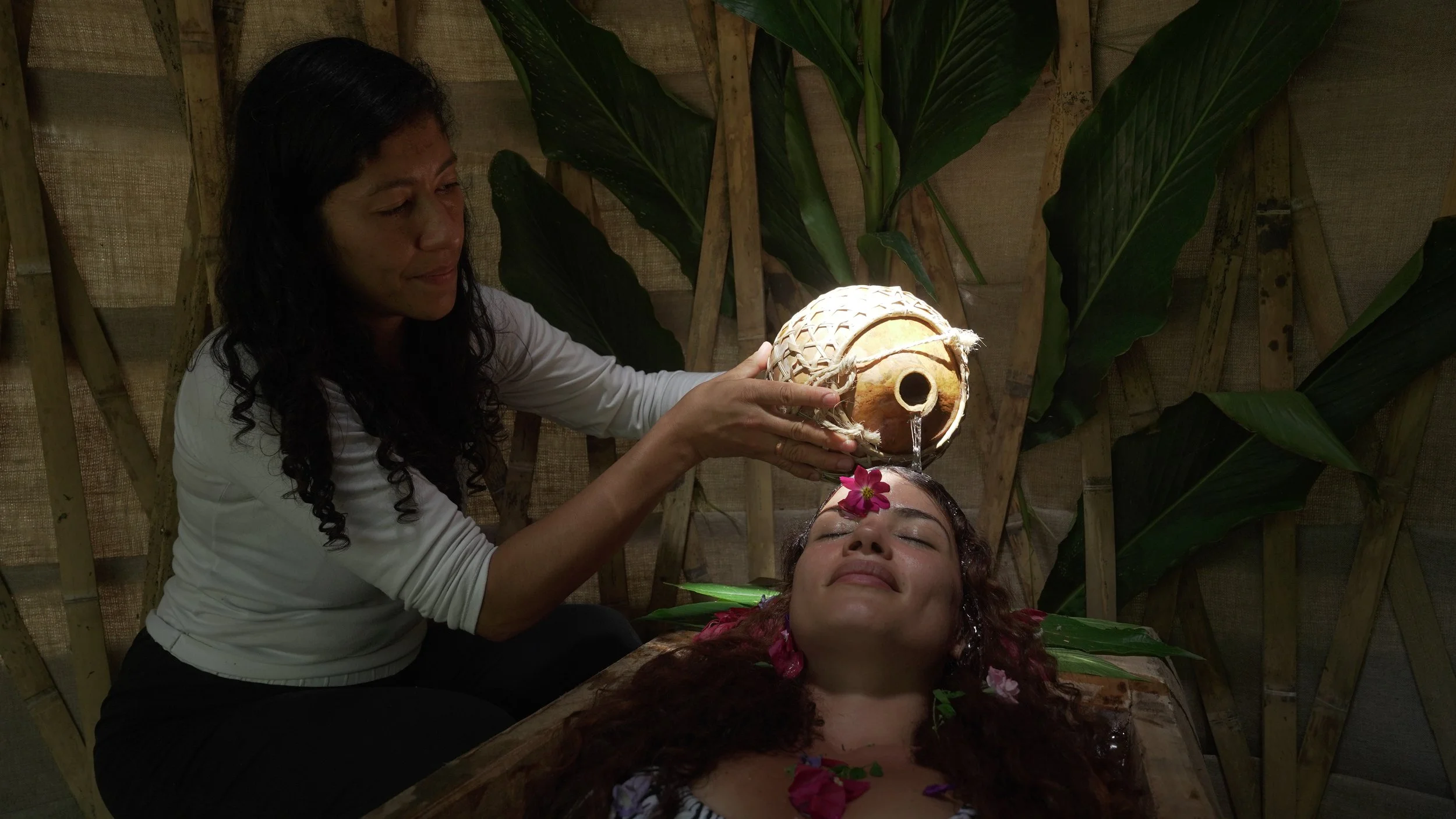 Una mujer recibe un masaje con agua del cotón en una ceremonia tradicional, mientras otra mujer está acostada con flores en el cabello en un ambiente natural con plantas grandes de fondo.