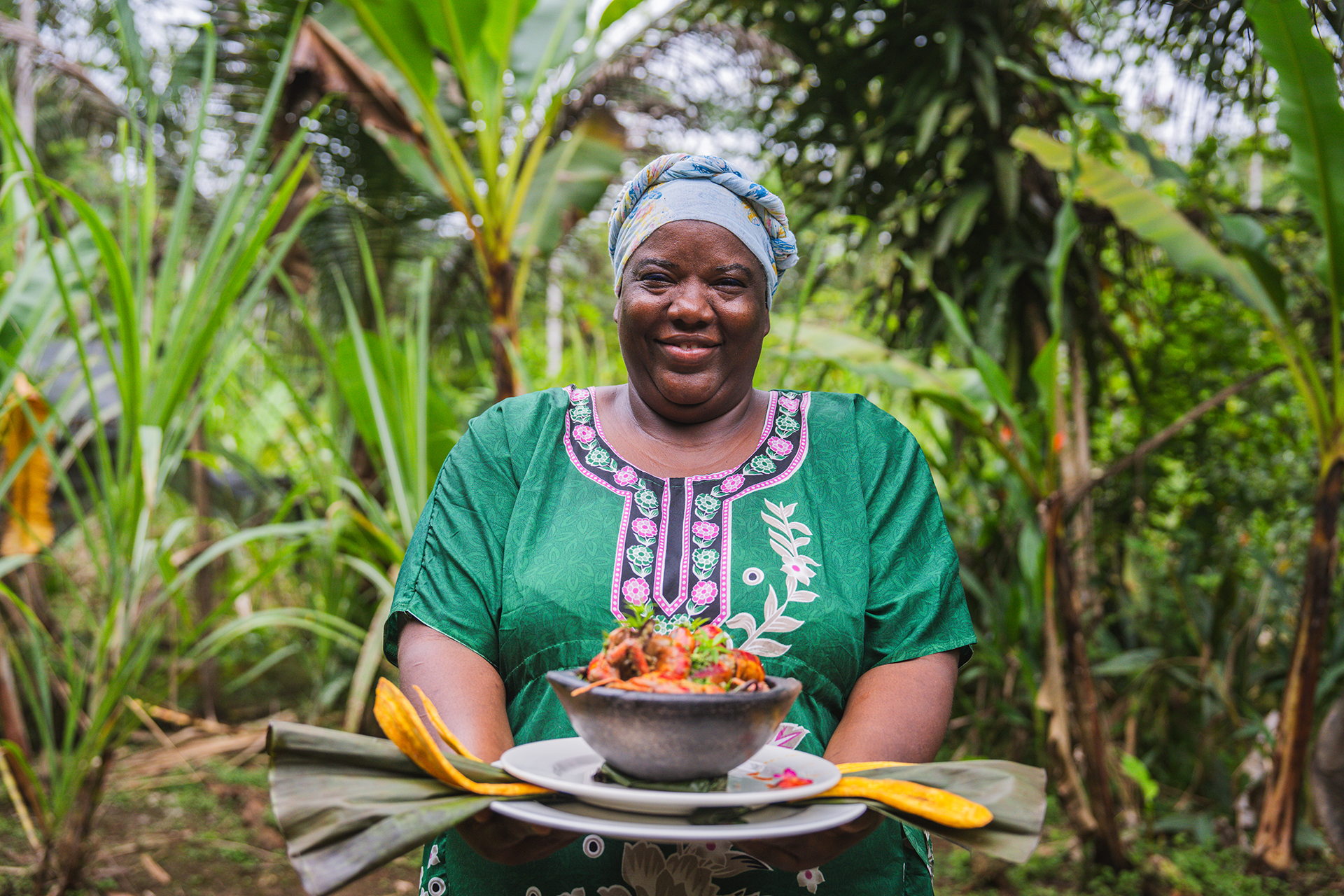 Mujer en un campo de cultivo sosteniendo un plato con comida tradicional, vestida con ropa elegante y un tocado en la cabeza, rodeada de vegetación