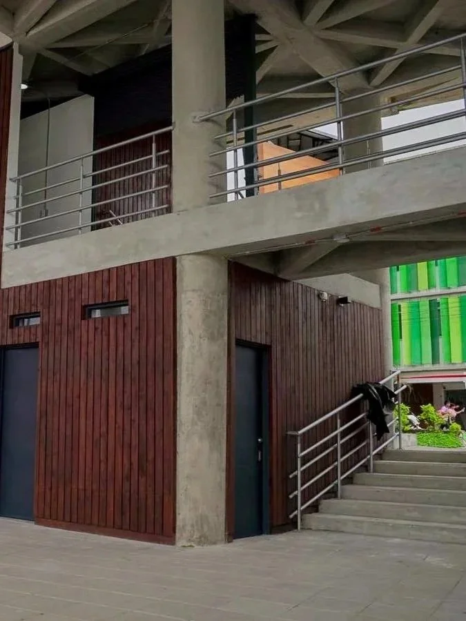 Estructura moderna con paredes de madera y concreto, con una escalera de granito y barandales metálicos, en un espacio interior o exterior con paredes de pantalla verde y decoración en el fondo.
