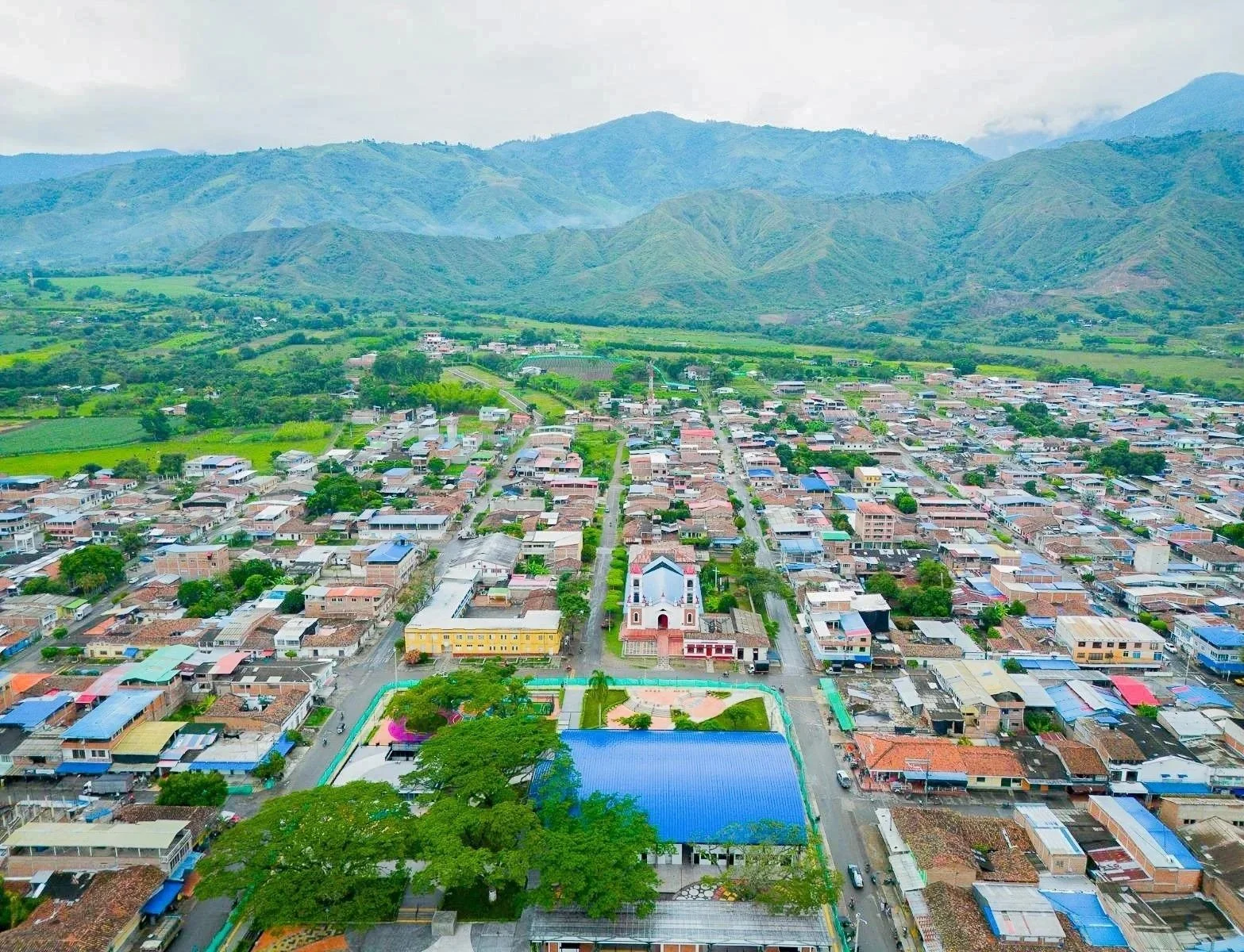 Vista aérea de un pueblo en un valle con montañas verdes en el fondo, incluyendo una iglesia blanca en el centro, rodeada de casas y edificios pequeños.
