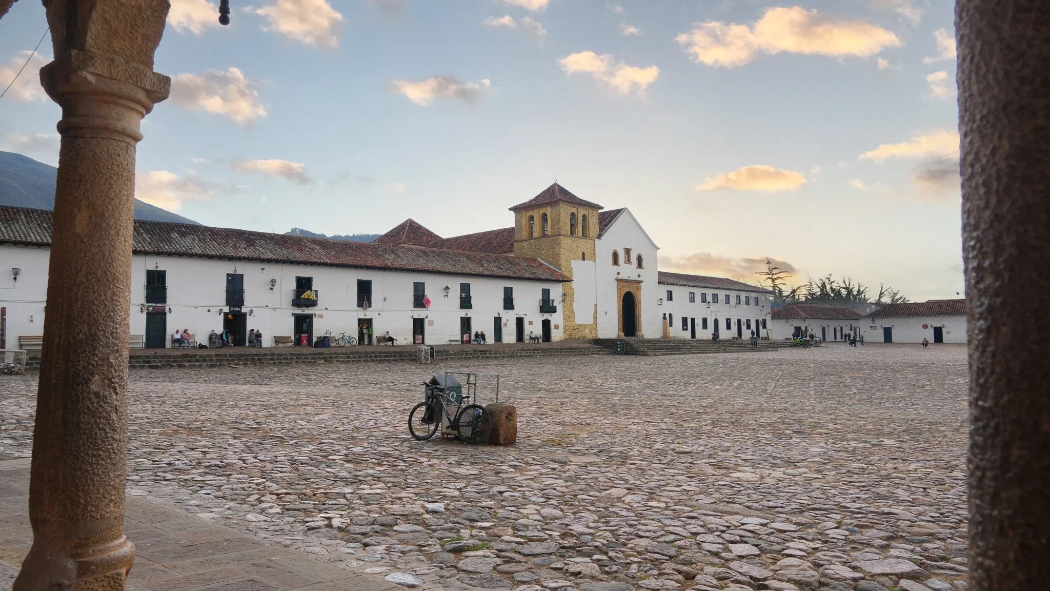 Plaza en un pueblo colonial con calles de piedra, edificios blancos y techo de tejas. Hay una iglesia con campanario, personas sentadas en bancos, bicicletas y árboles.