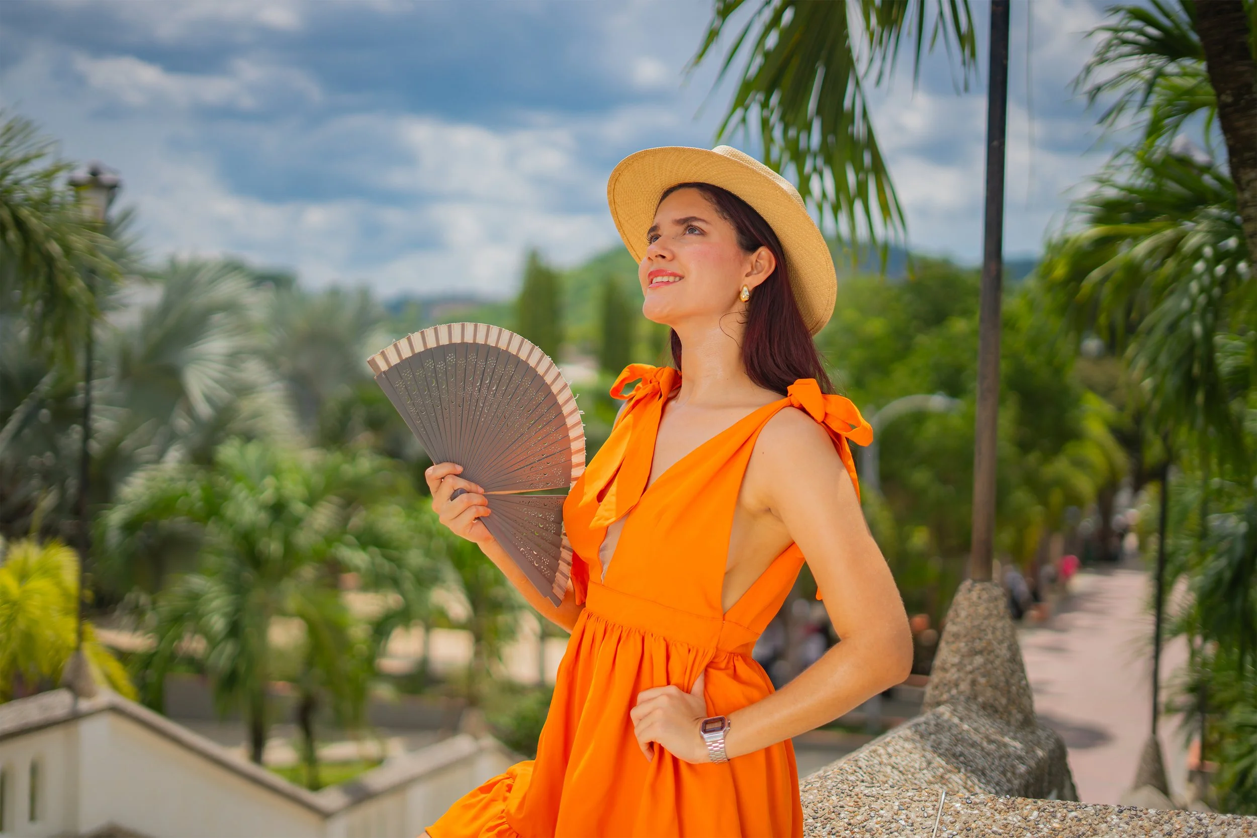 Mujer con vestido naranja y sombrero de paja sostiene un abanico en un parque con árboles y cielo nublado.