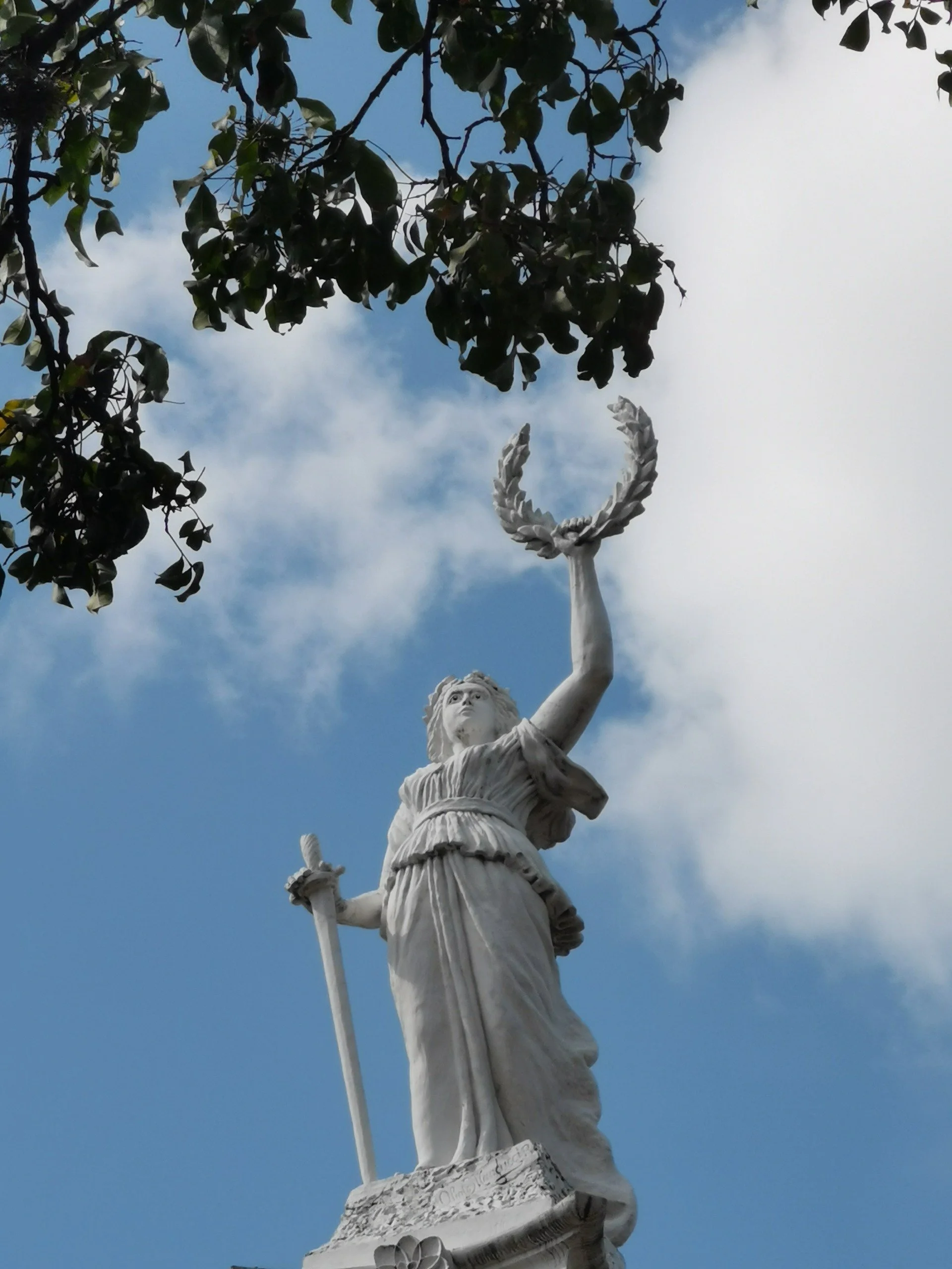 Escultura de una figura femenina con una corona de laurel en la mano derecha, sobre un pedestal decorado, contra un cielo con nubes.