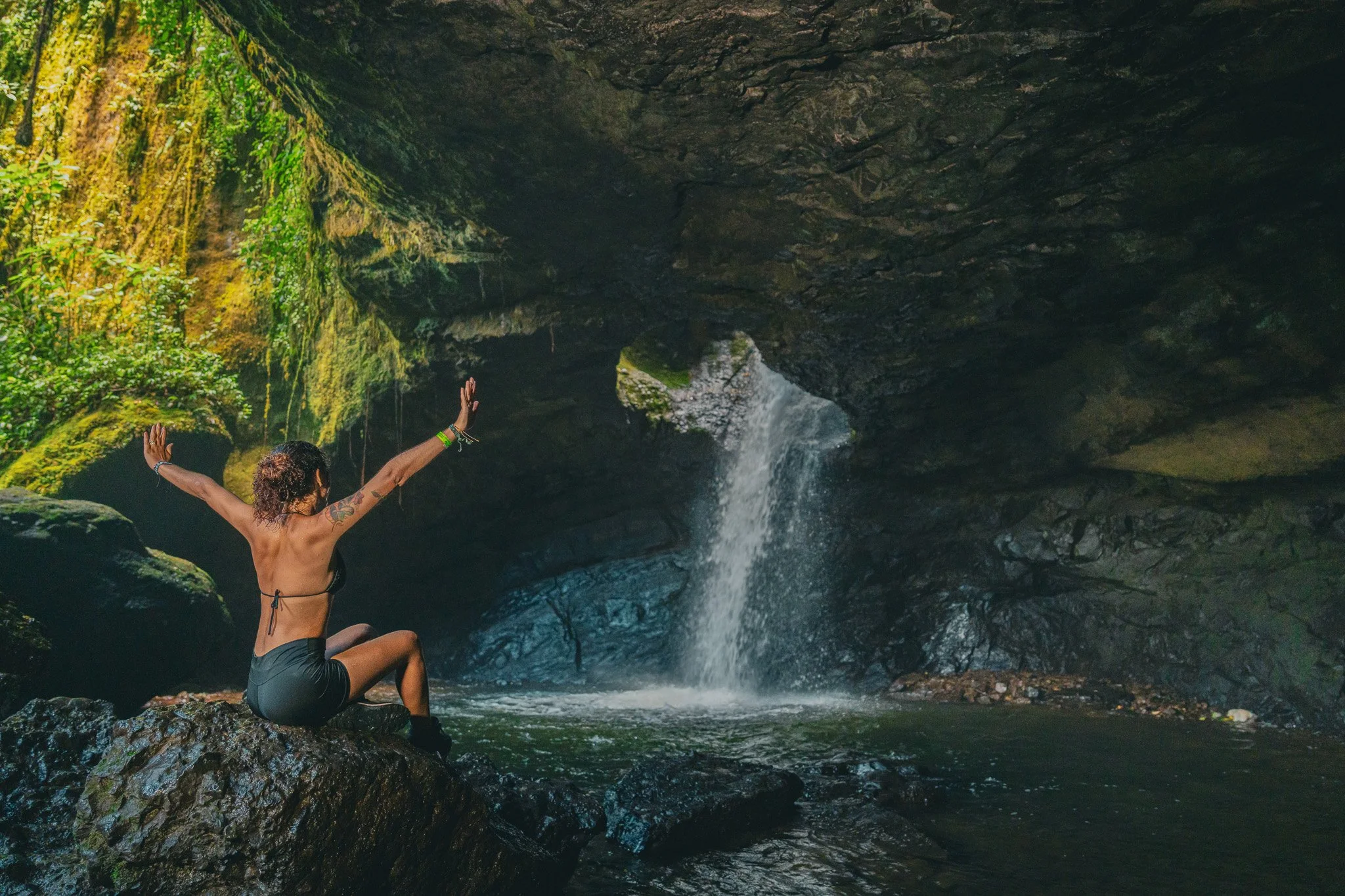 Persona en bikini sentado en roca dentro de una cueva con cascada, agua cayendo de la parte superior.