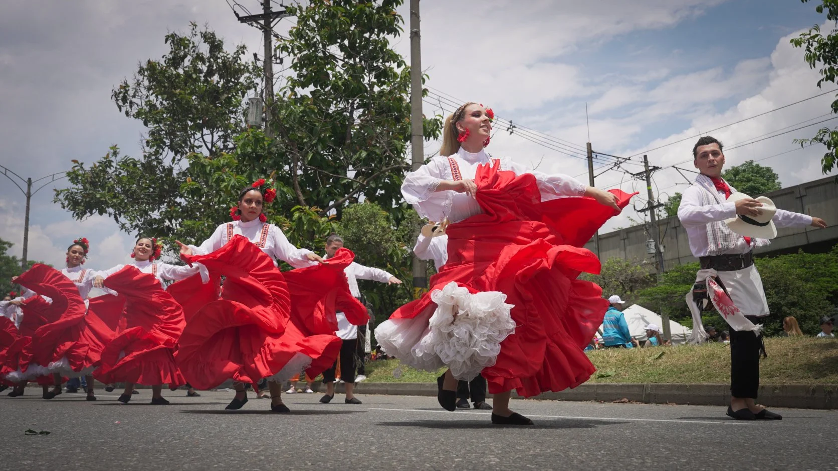 Grupo de personas bailando en una celebración folklórica, vestidas con trajes tradicionales color rojo y blanco, en una calle con árboles, cables y un muro de fondo.