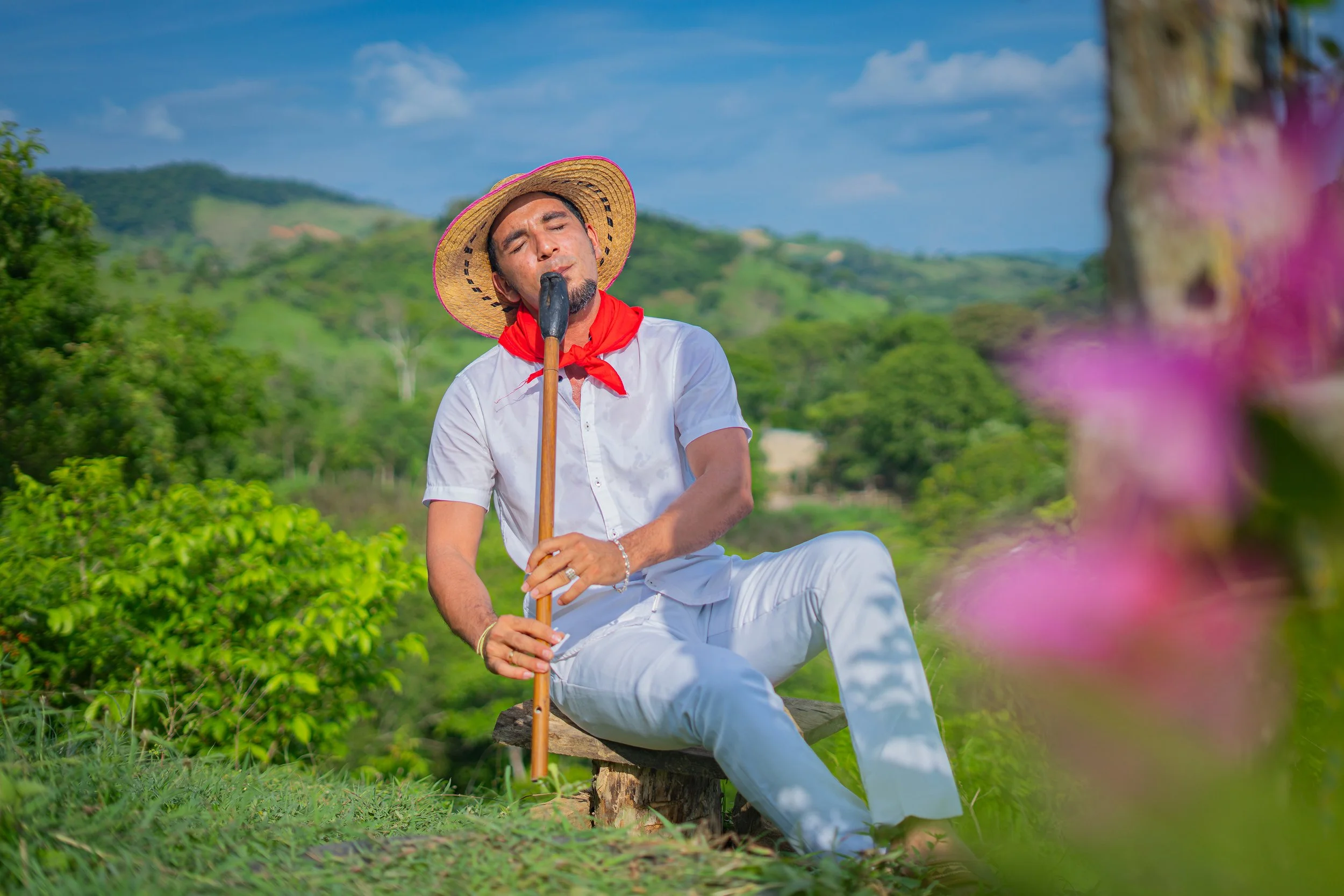 Hombre vestido de blanco, con sombrero de paja y pañuelo rojo, sentado en un banco de madera en un campo verde, con colinas y cielo azul al fondo, sosteniendo un bastón.