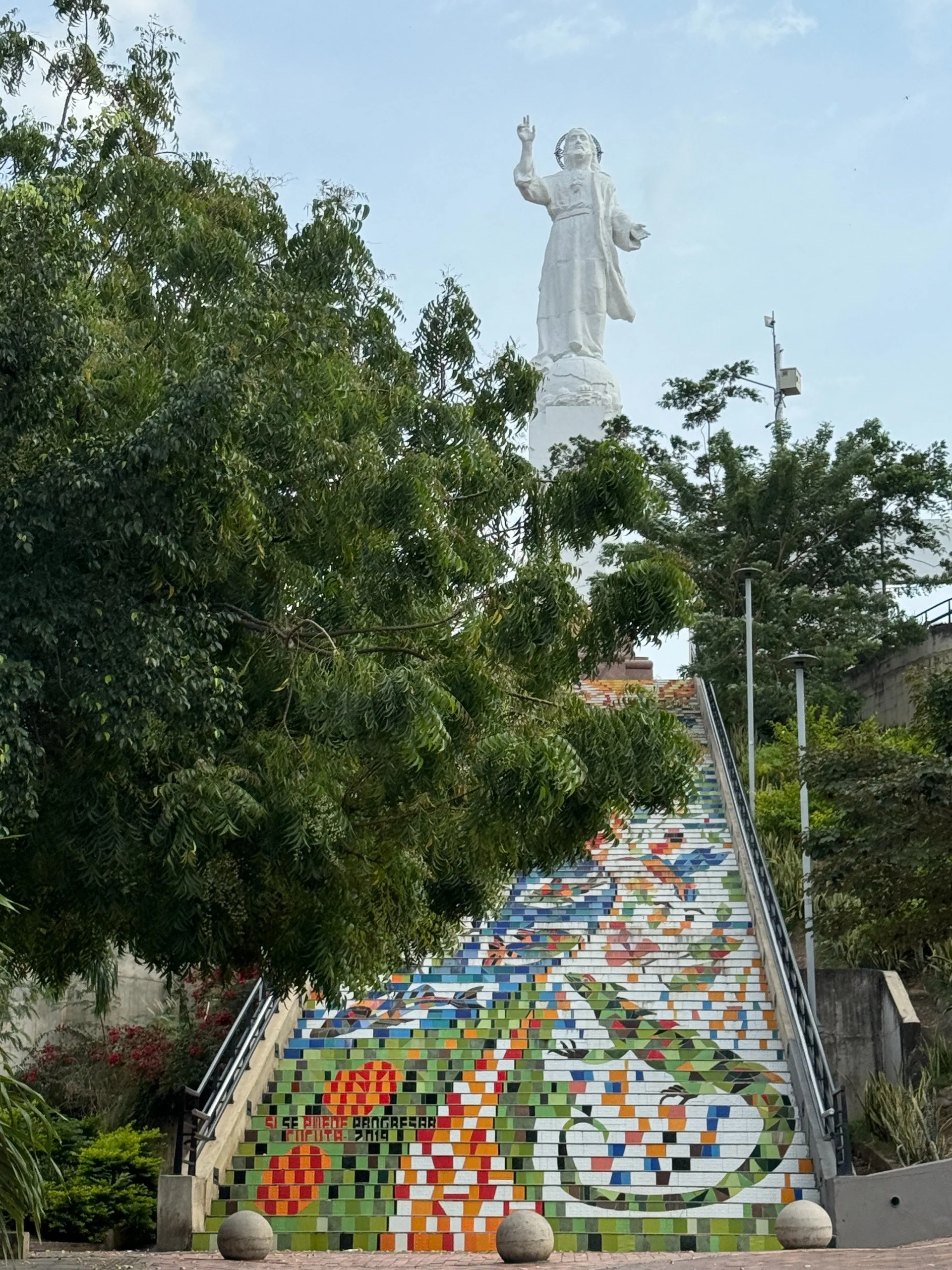 Escaleras decoradas con mosaicos de colores y un mural con abejas en la parte inferior, rodeadas de árboles y vegetación.