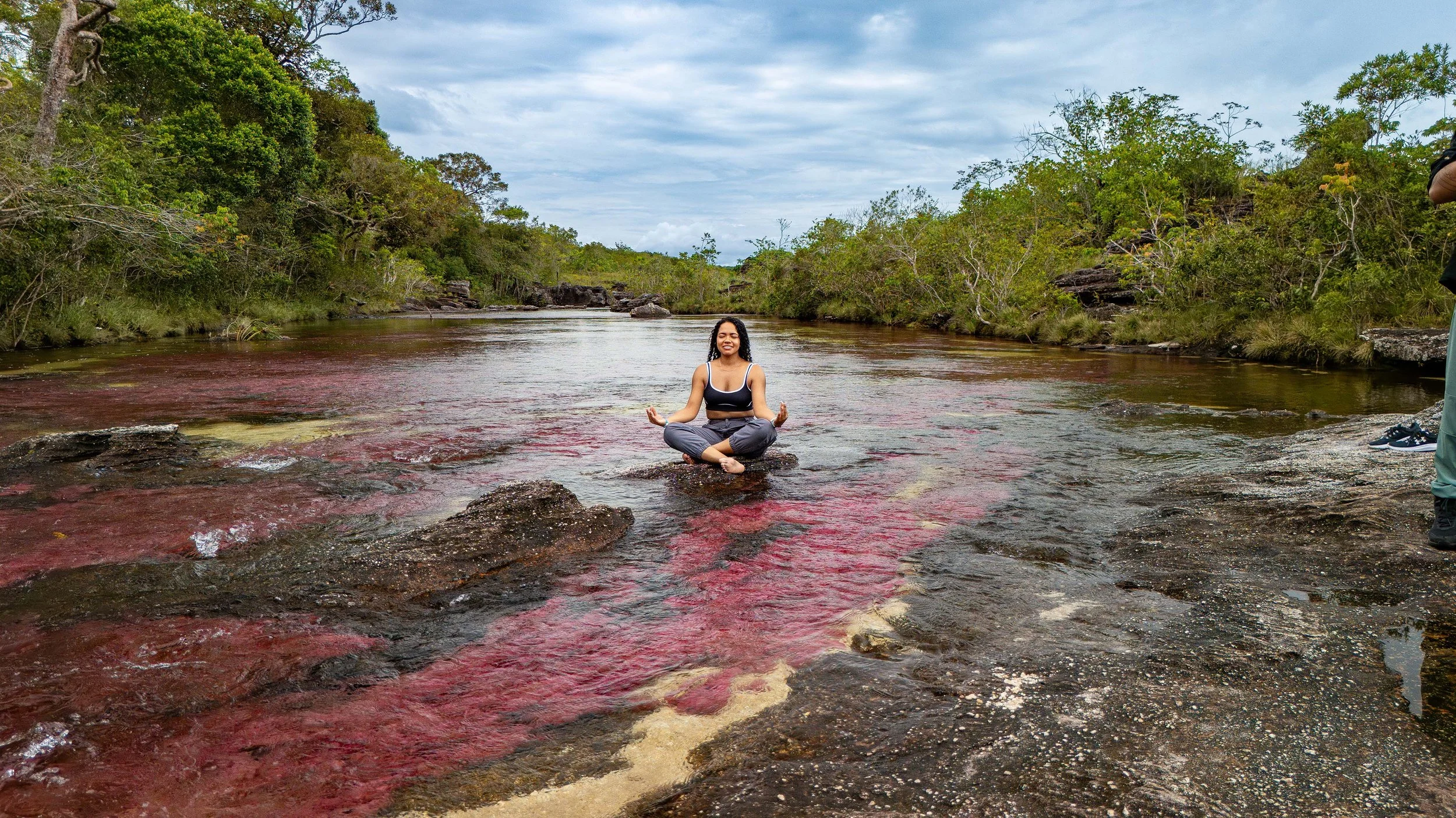 Mujer practicando yoga o meditación en una roca en medio de un río rodeada de vegetación y árboles, con cielo nublado.