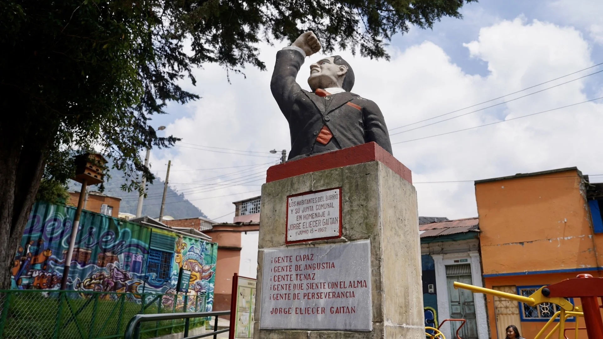 Escultura de un hombre con el puño levantado, ubicada en una plaza en un barrio popular, con casas coloridas y grafiti en las paredes, cielo nublado y montañas al fondo.