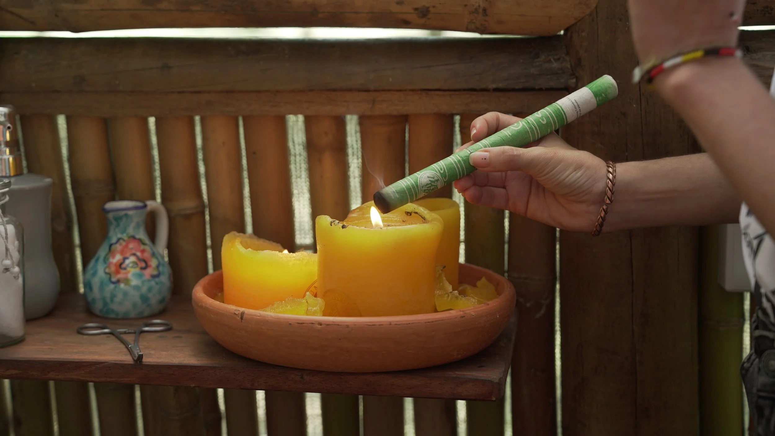 Mano colocando una vela en una vela amarilla en una mesa de madera con jarrones de cerámica y un fondo de paredes de madera.