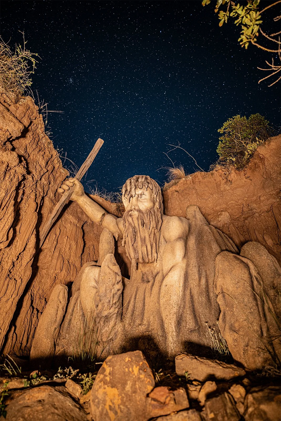 Escultura de Jesucristo con una cruz tallada en piedra, en un entorno rocoso bajo un cielo estrellado.