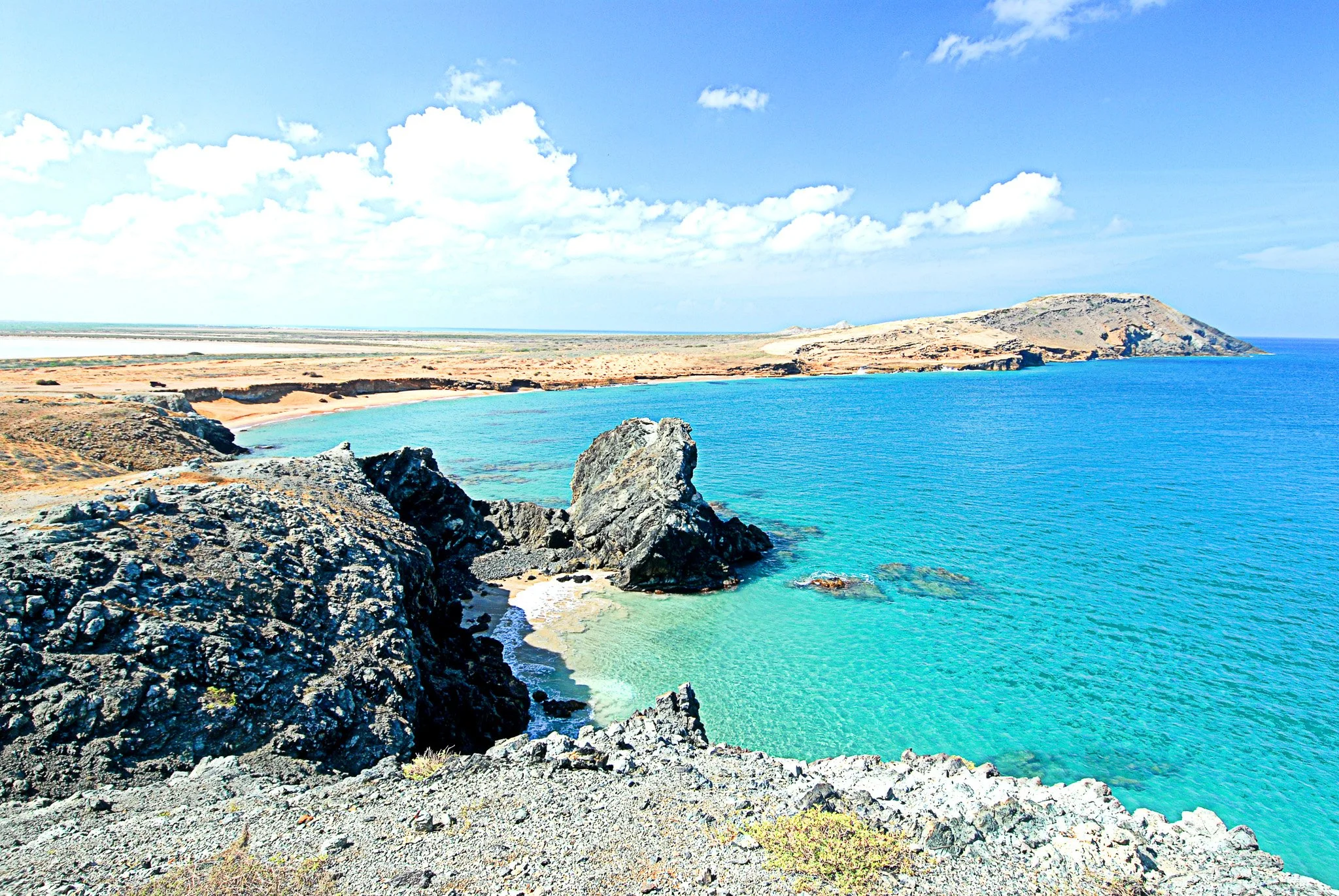Costa de un mar azul claro con rocas negras en primer plano y una playa de arena, con colinas y cielo despejado con algunas nubes.