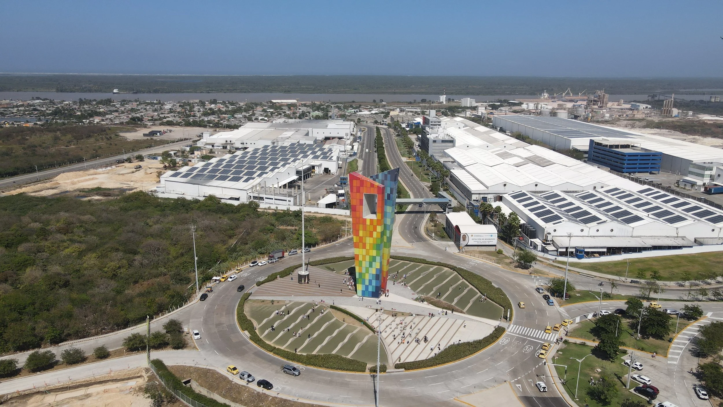 Vista aérea de un complejo industrial con techos de paneles solares, calles con vehículos y un monumento colorido en primer plano en una ciudad cerca de un río o cuerpo de agua.