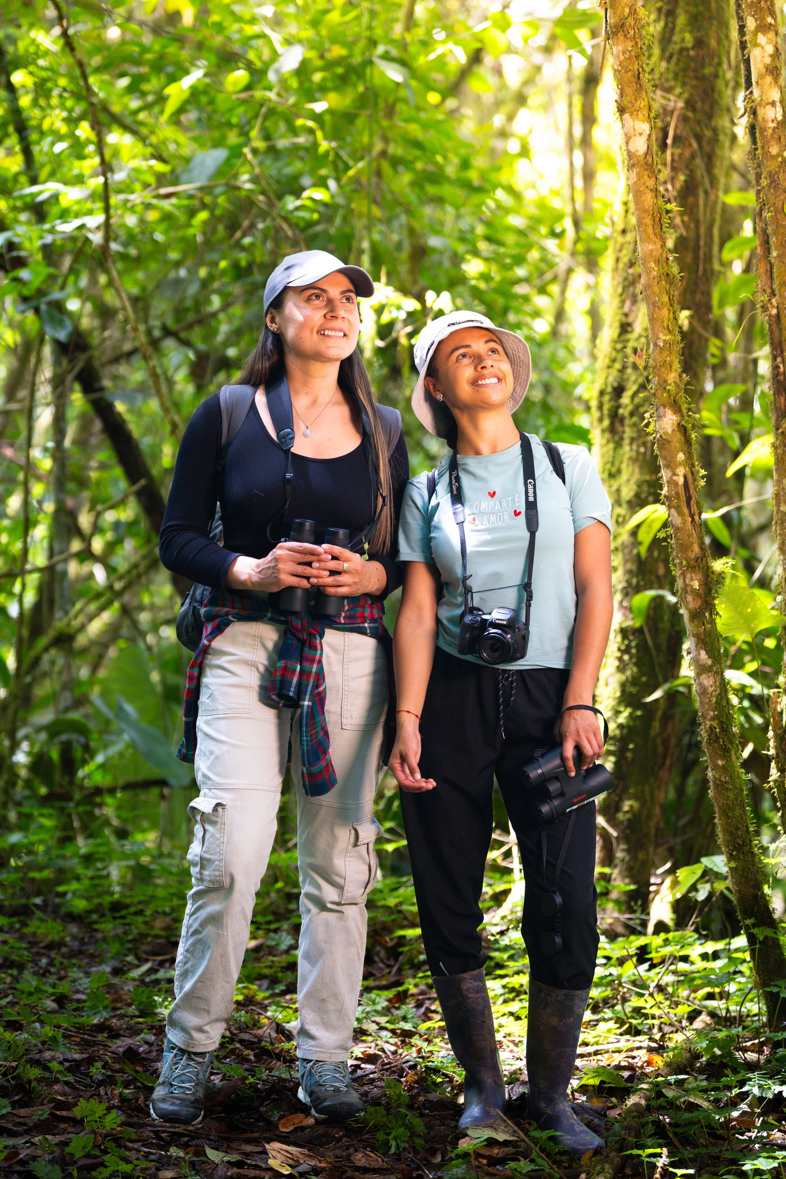 Dos mujeres en ropa de senderismo observando la naturaleza en una selva densa, una con binoculares y la otra con cámara.