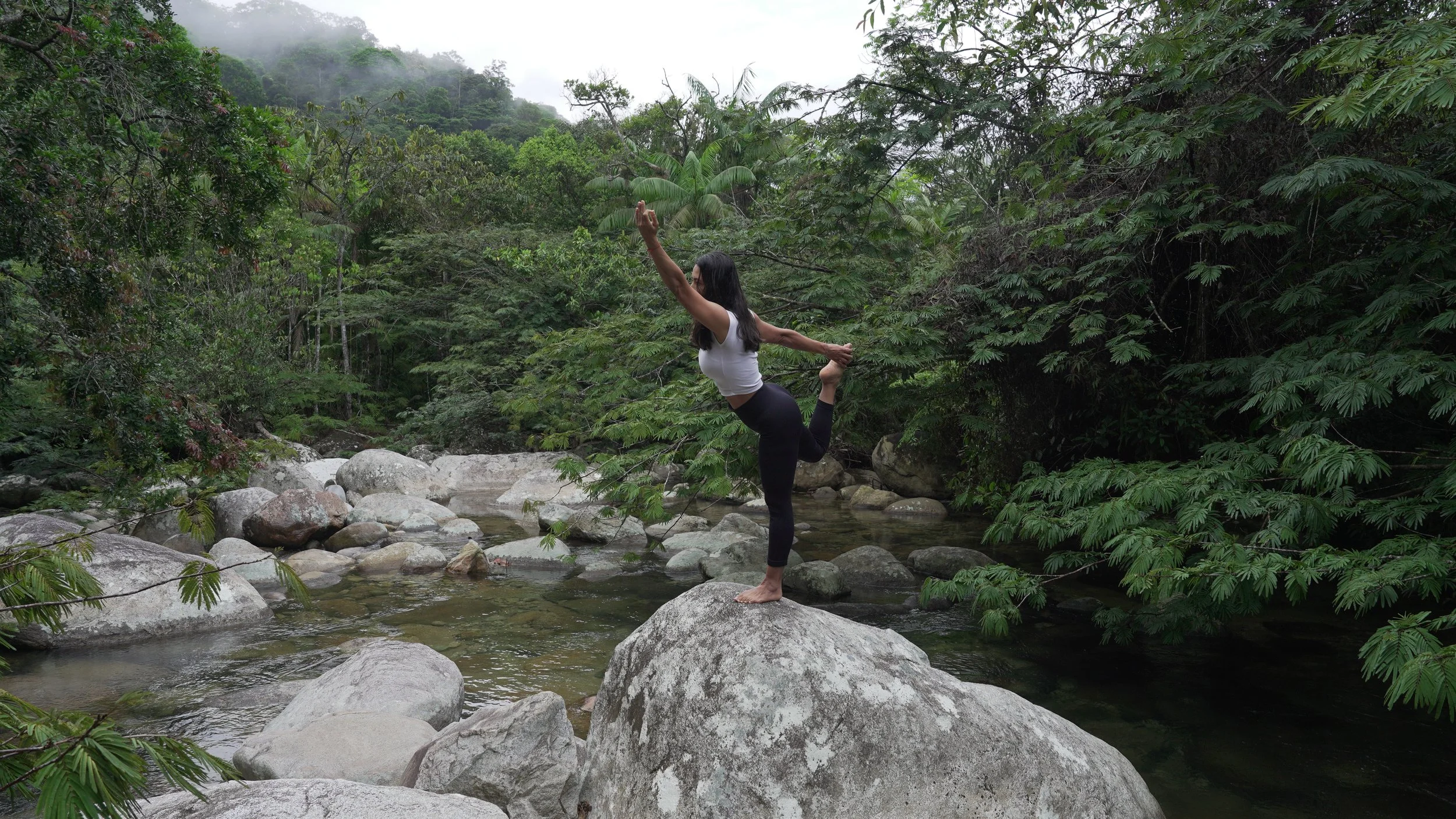 Mujer practicando yoga en una roca en un río rodeado de vegetación densa, en un entorno natural y lluvioso.