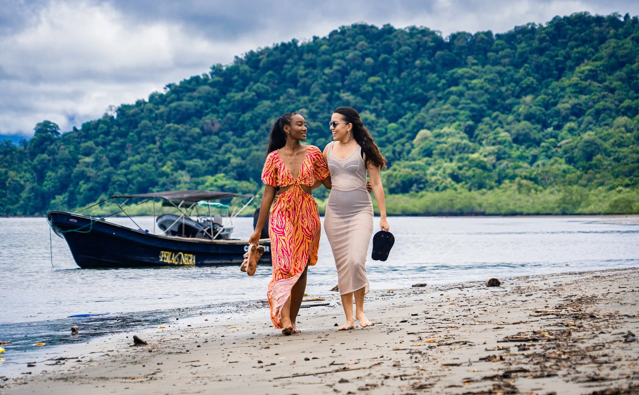 Dos mujeres caminando juntas por la playa con un bote de fondo, rodeadas de una región montañosa y un cielo nublado.