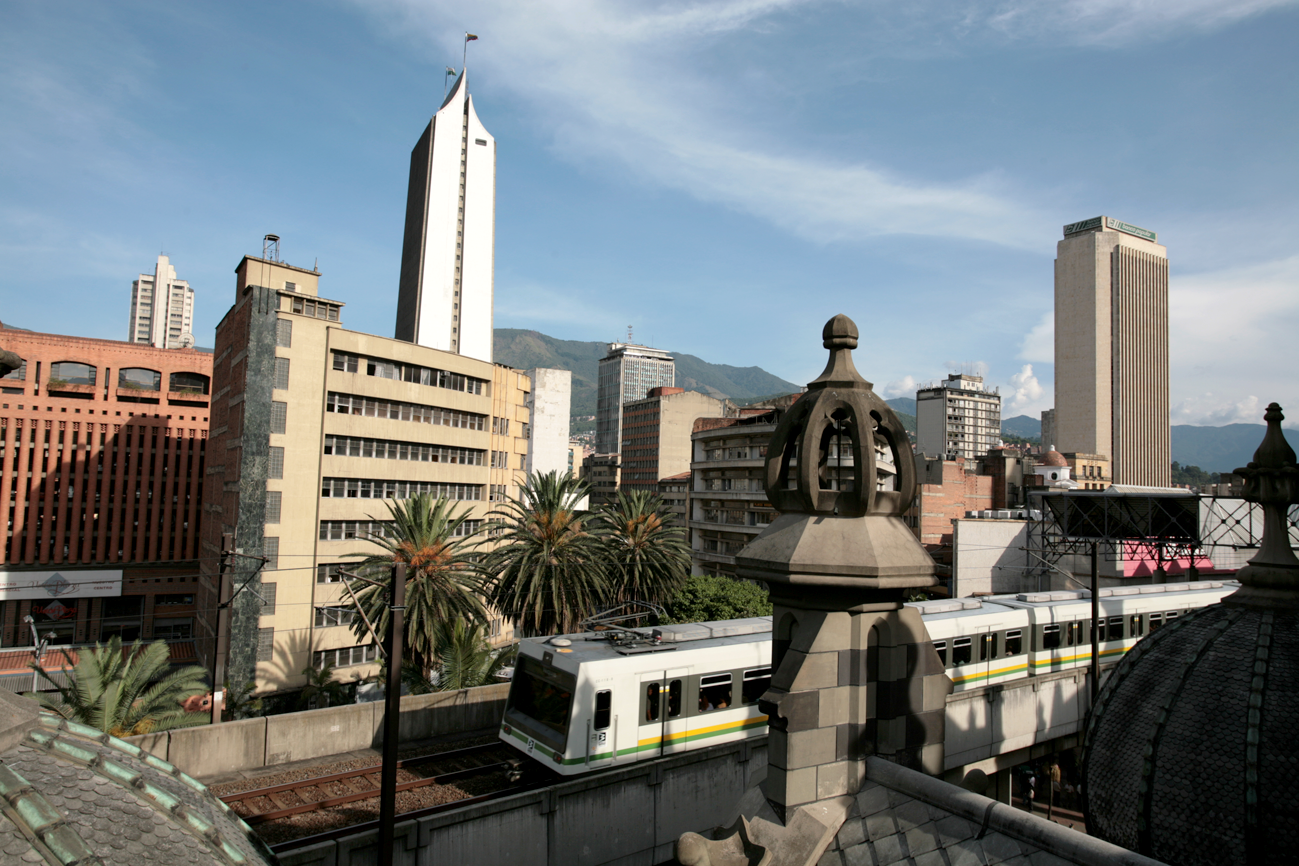 Vista de la ciudad con edificios altos, un tren en la vía de tren, palmeras, y montañas al fondo, en un día soleado.
