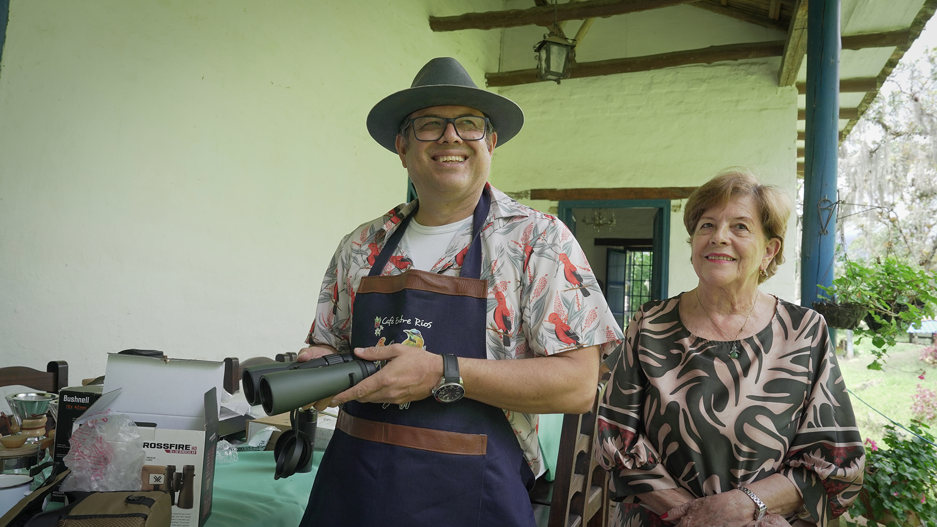 Dos personas, un hombre con sombrero y gafas, y una mujer, posan juntas en un ambiente de interior con exteriores visibles. Están sonriendo y parecen estar en una reunión o evento, con objetos y equipo en una mesa frente a ellos.