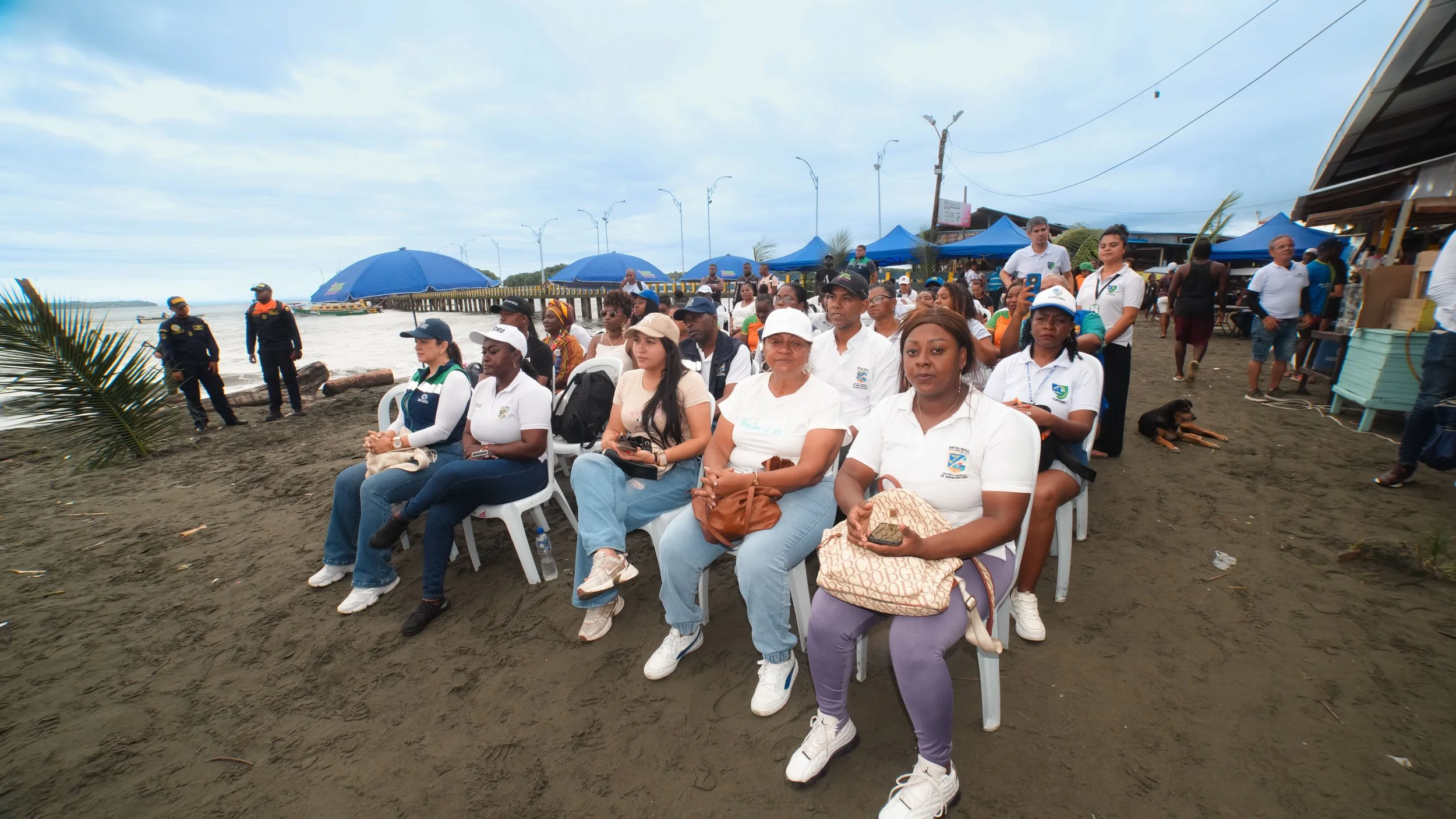 grupo de personas asistiendo a un evento en la playa, algunas sentadas en sillas de plástico, con paraguas y sombreros, mientras otras están de pie, fondos con toldos azules y estructura junto al mar.