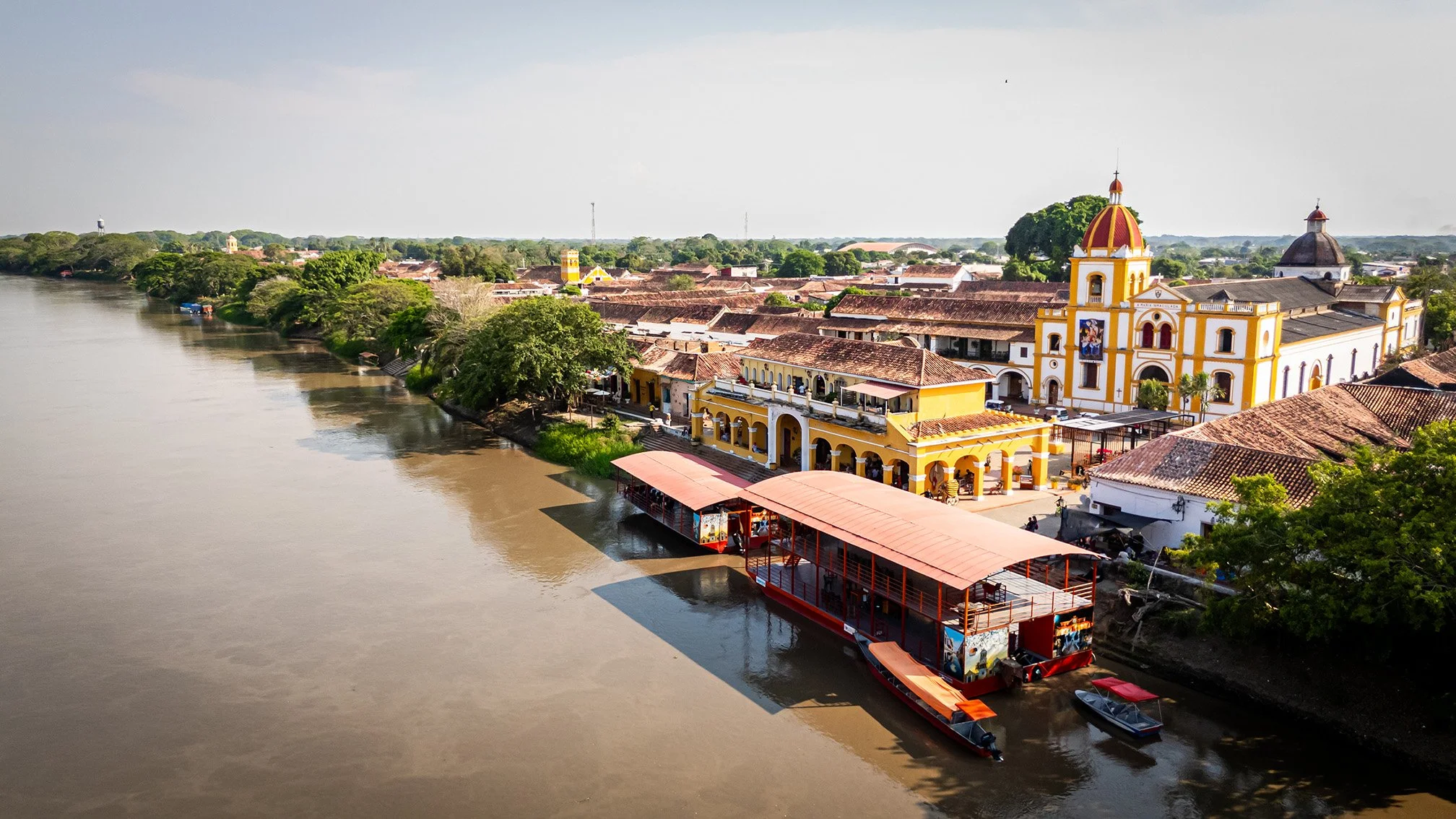 Vista aérea de un río con un pueblo a la orilla, resaltando una iglesia colonial amarilla con cúpula roja y un techo de tejas rojas, y un muelle con una lancha turística.