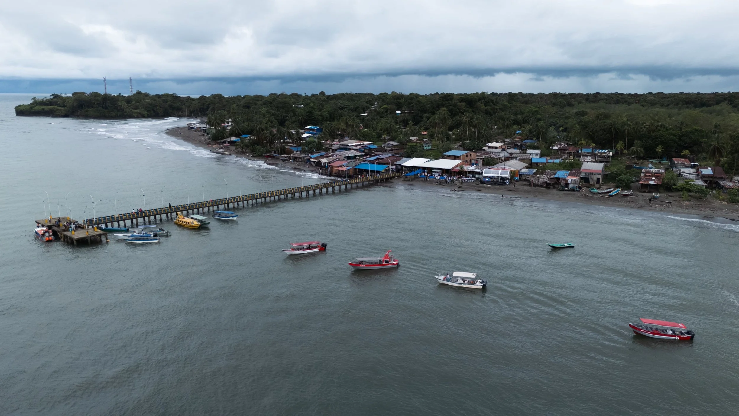 Vista aérea de un pueblo costero con un muelle, varias embarcaciones en el agua y una zona boscosa con nubes oscuras en el cielo.