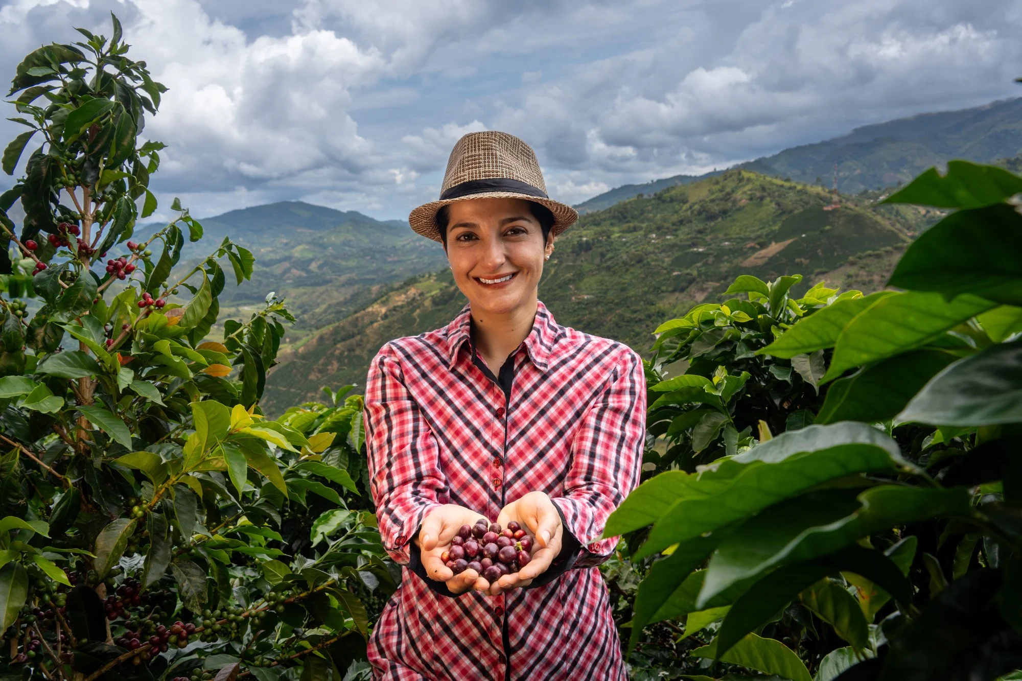 Mujer sonriendo en un cultivo de cafetales, sosteniendo bebidas de café en sus manos, con montañas y cielo nublado al fondo.