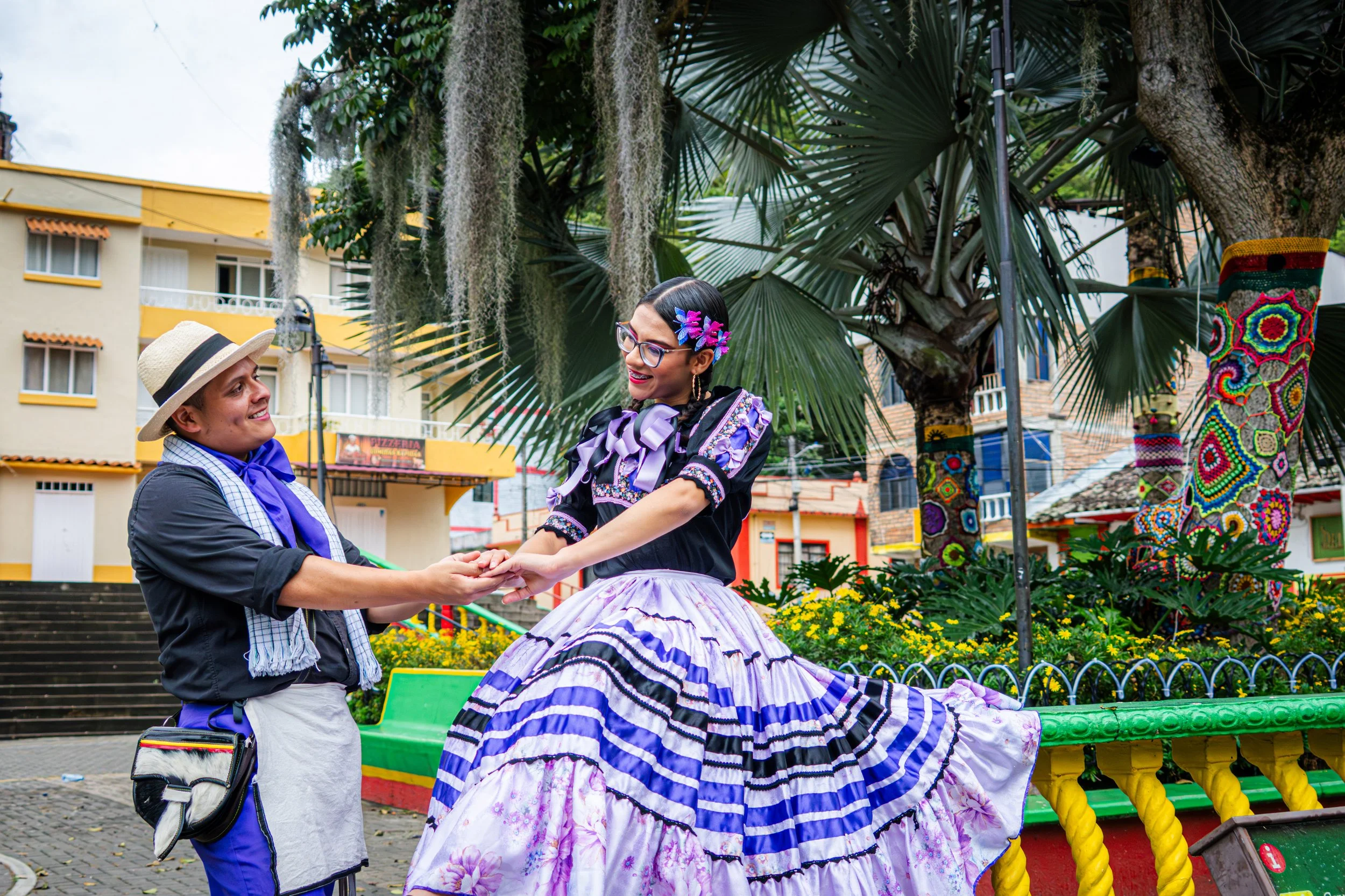 Hombre y mujer bailando en un parque con vegetación y edificios de fondo, ella viste un vestido tradicional colorido y él lleva sombrero y pañuelo azul.