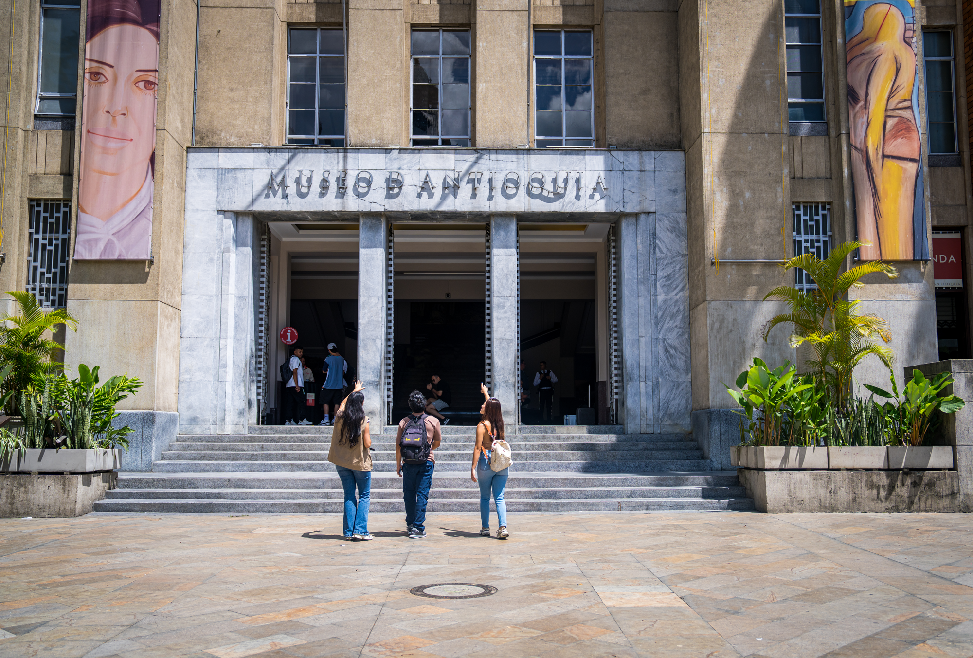 Fachada del Museo de Antioquia con señalización en letras metálicas y dos mosaicos con retratos de personas en las paredes. Estudiantes conversando en la entrada y plantas decorativas en los costados.