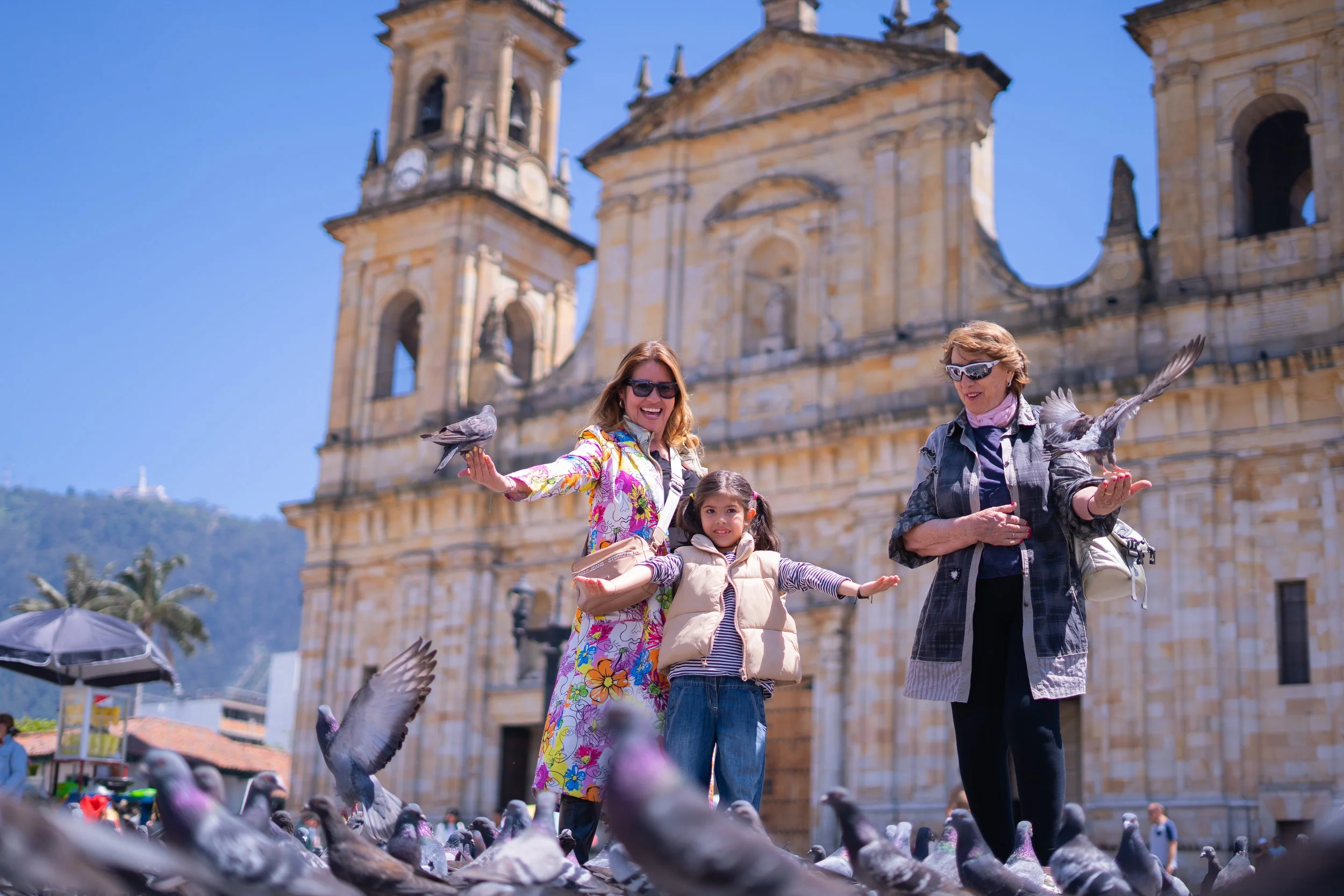 Tres mujeres y una niña posan en una plaza con una iglesia antigua de fondo. Una de las mujeres tiene gafas oscuras y una blusa de colores, otra mujer usa gafas y una chaqueta de cuadros, la niña luce una chaqueta beige y una camiseta a rayas. Todas están rodeadas de palomas y parecen disfrutar del día soleado.