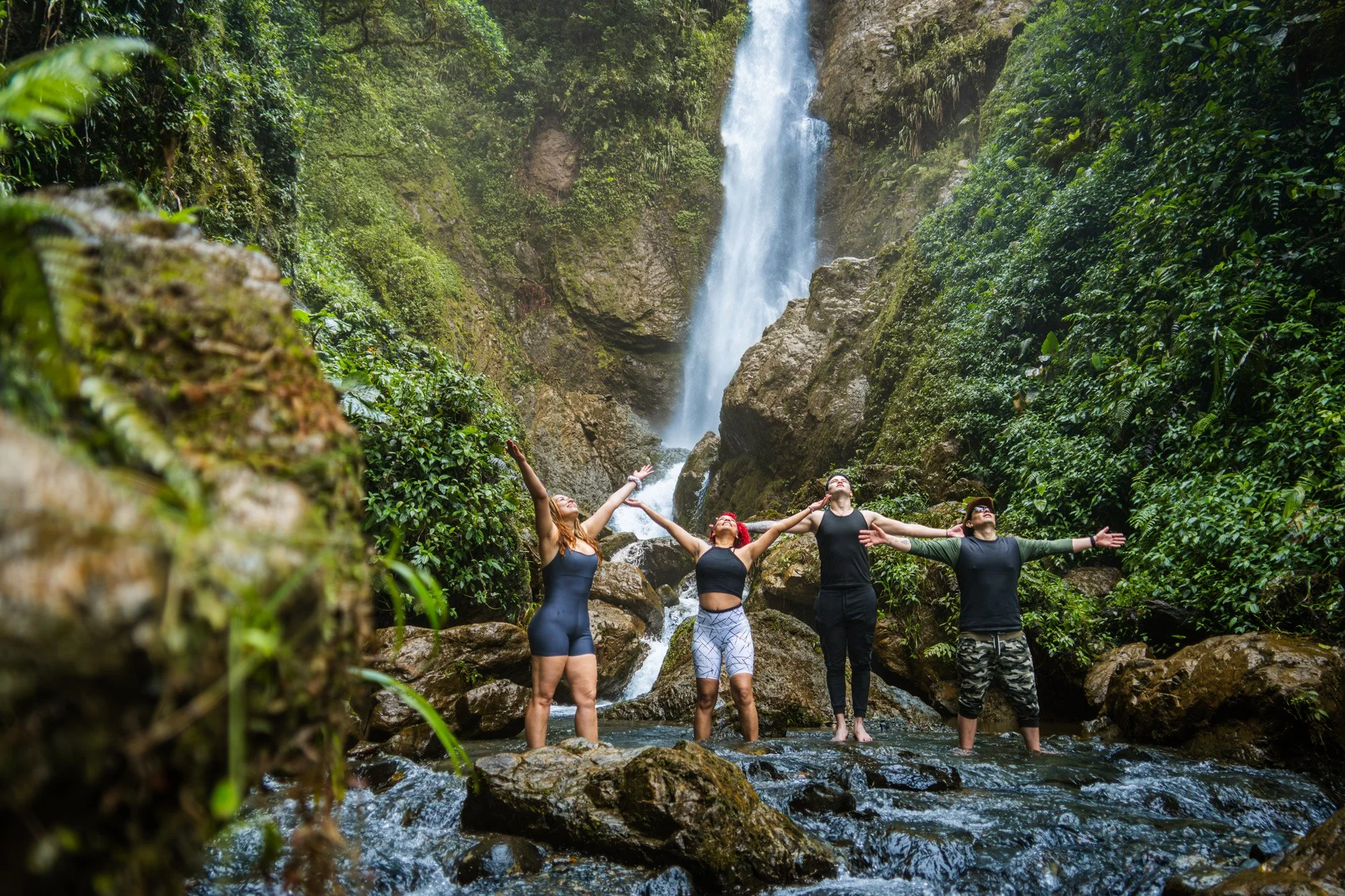Un grupo de cinco personas en ropa deportiva, de espaldas, con los brazos levantados en señal de alegría, en un río rodeado de vegetación verde y una cascada en el fondo.