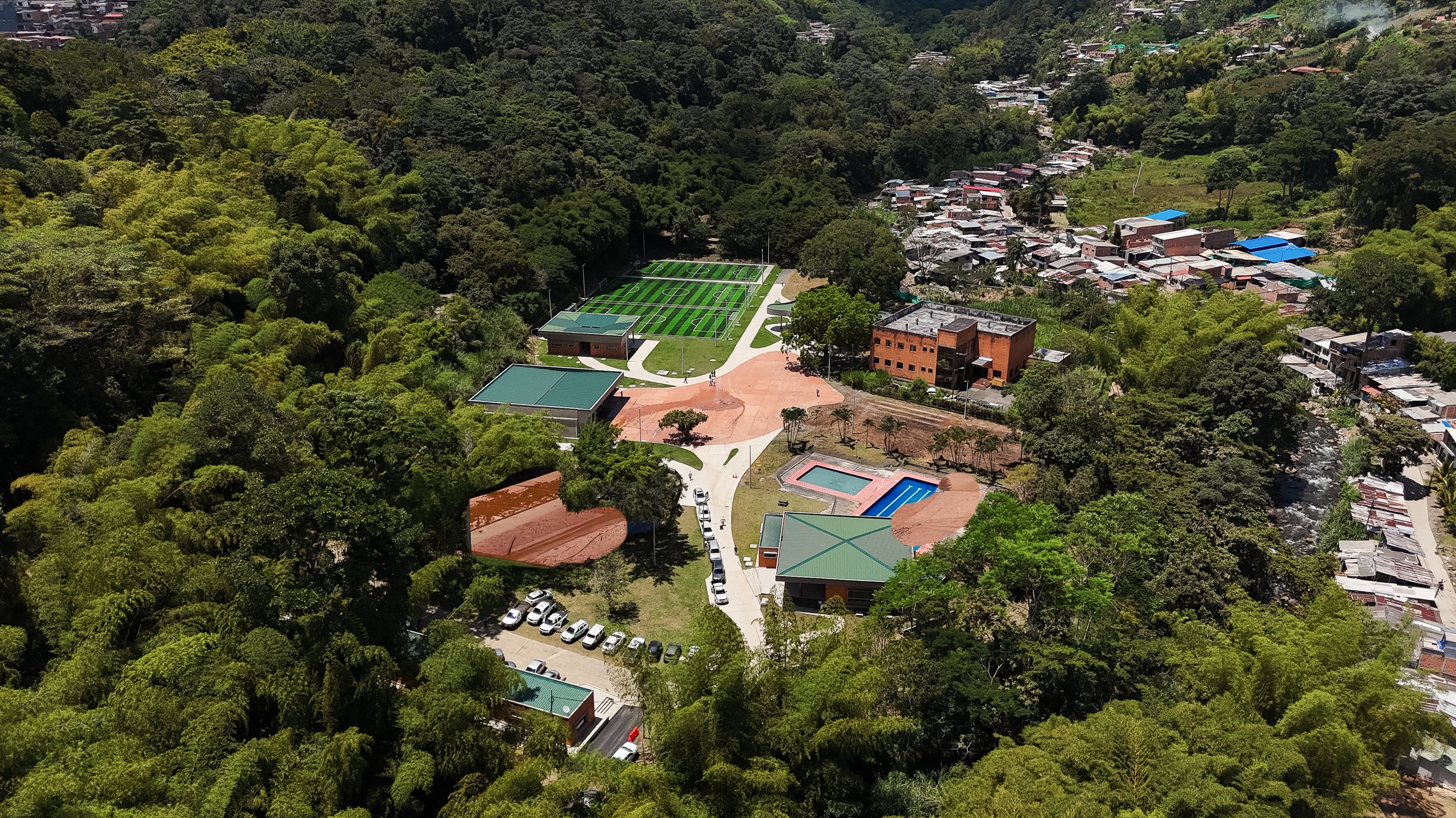 Vista aérea de un área recreativa rodeada de vegetación, con canchas de fútbol, piscina y caminos pavimentados en un entorno montañoso y residencial.