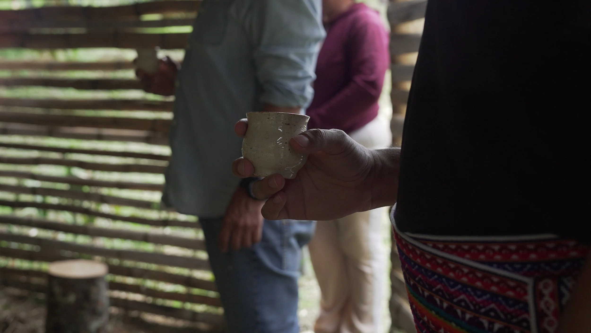 Persona sosteniendo una taza de barro en un entorno rústico con paredes de madera y otras personas en el fondo.