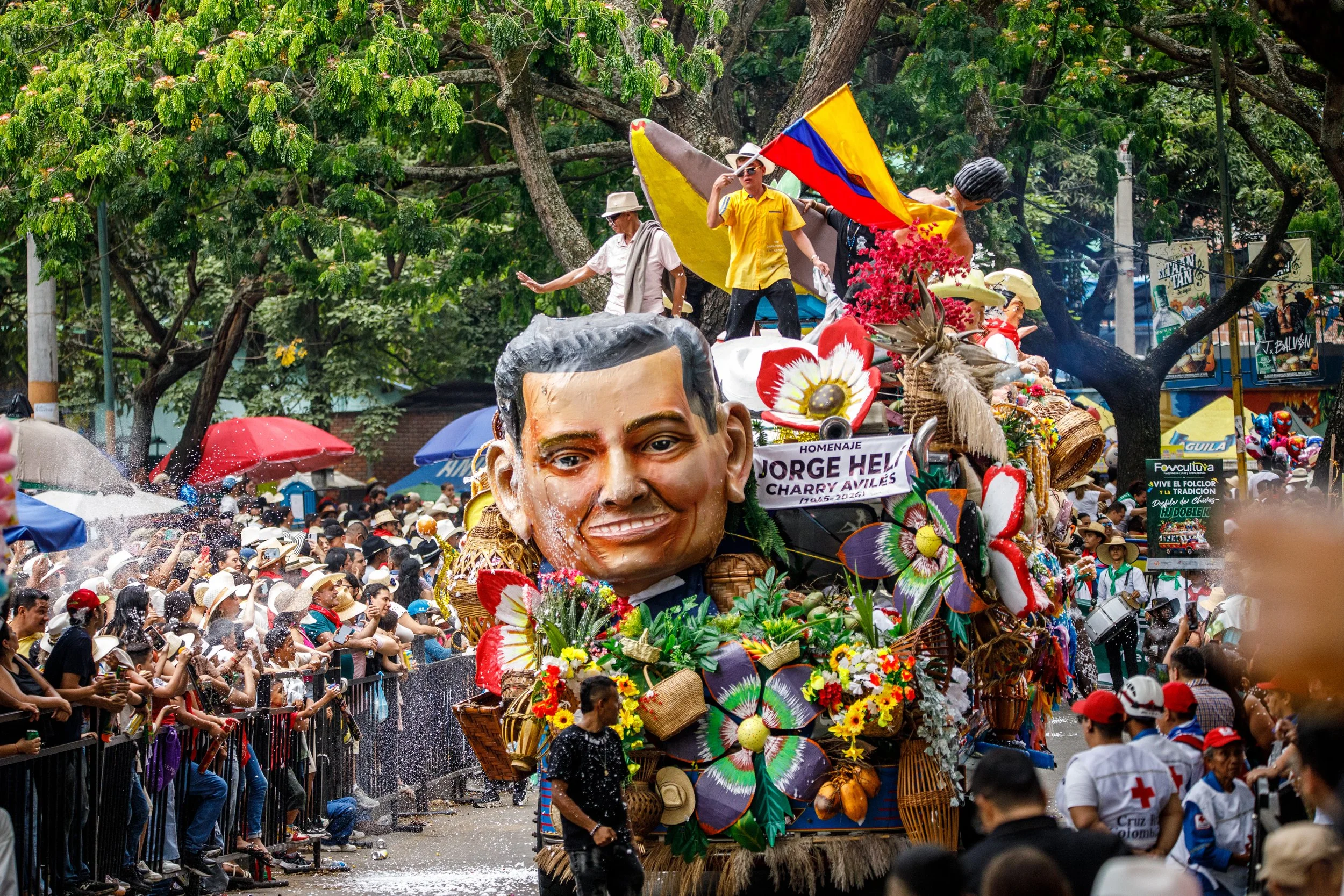 Personas en desfile con un carro alegórico decorado con figuras y flores, durante una celebración en un espacio público rodeado de árboles y asistentes con sombreros, en un día soleado.