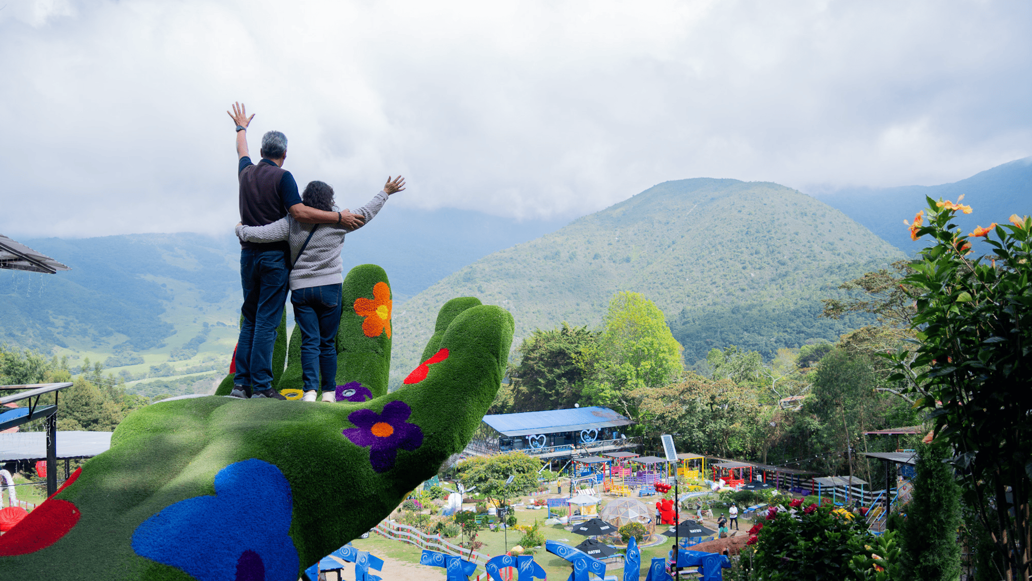 Tres personas, dos adultos y un niño, en la cima de una escultura de mano decorada con flores y colores vivos, con un paisaje montañoso y un parque de diversiones en el fondo.