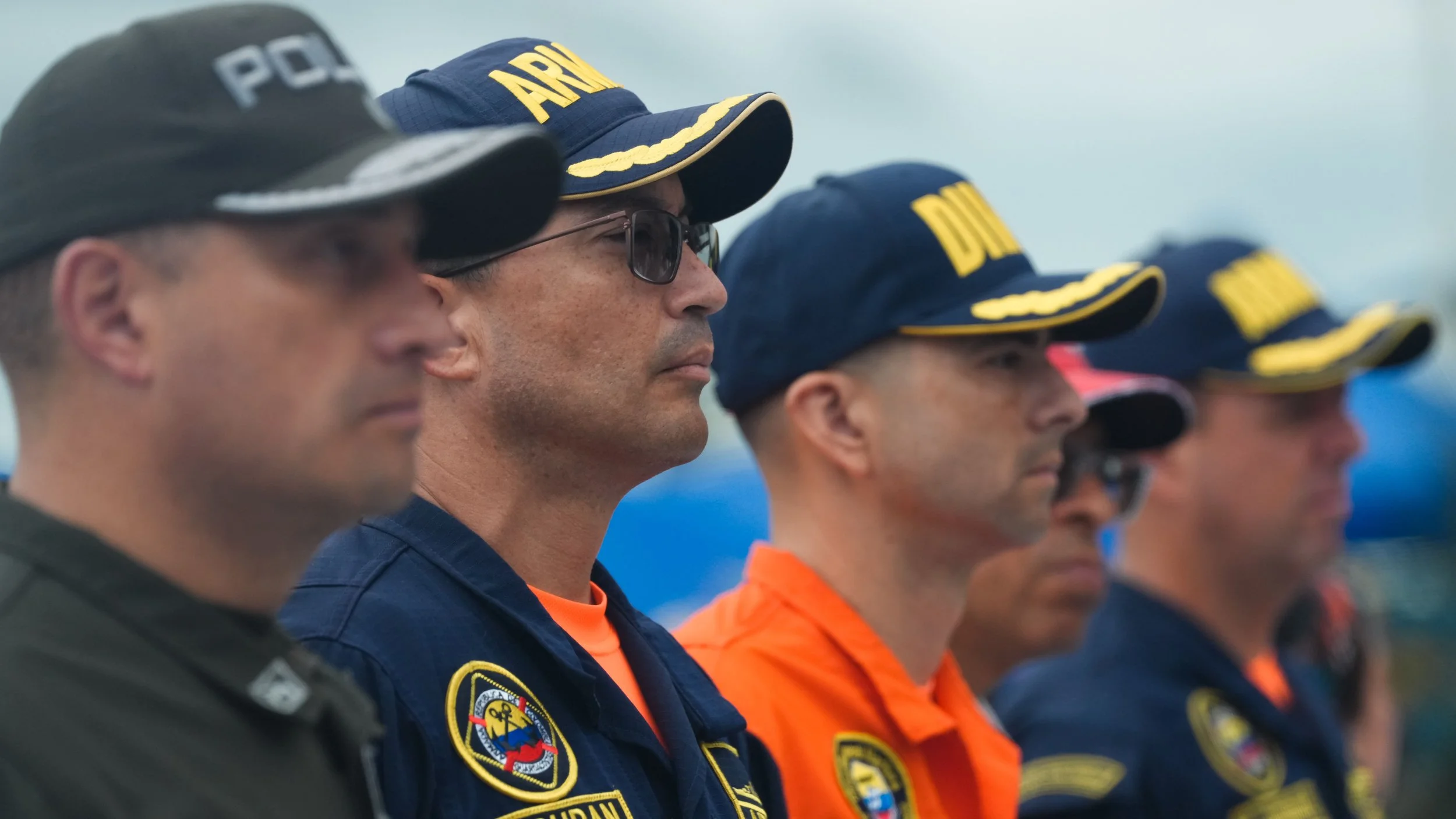 Grupo de oficiales de policía y personal de rescate en línea de formación, algunos con gorros y uniformes con insignias, observando atentamente hacia adelante.