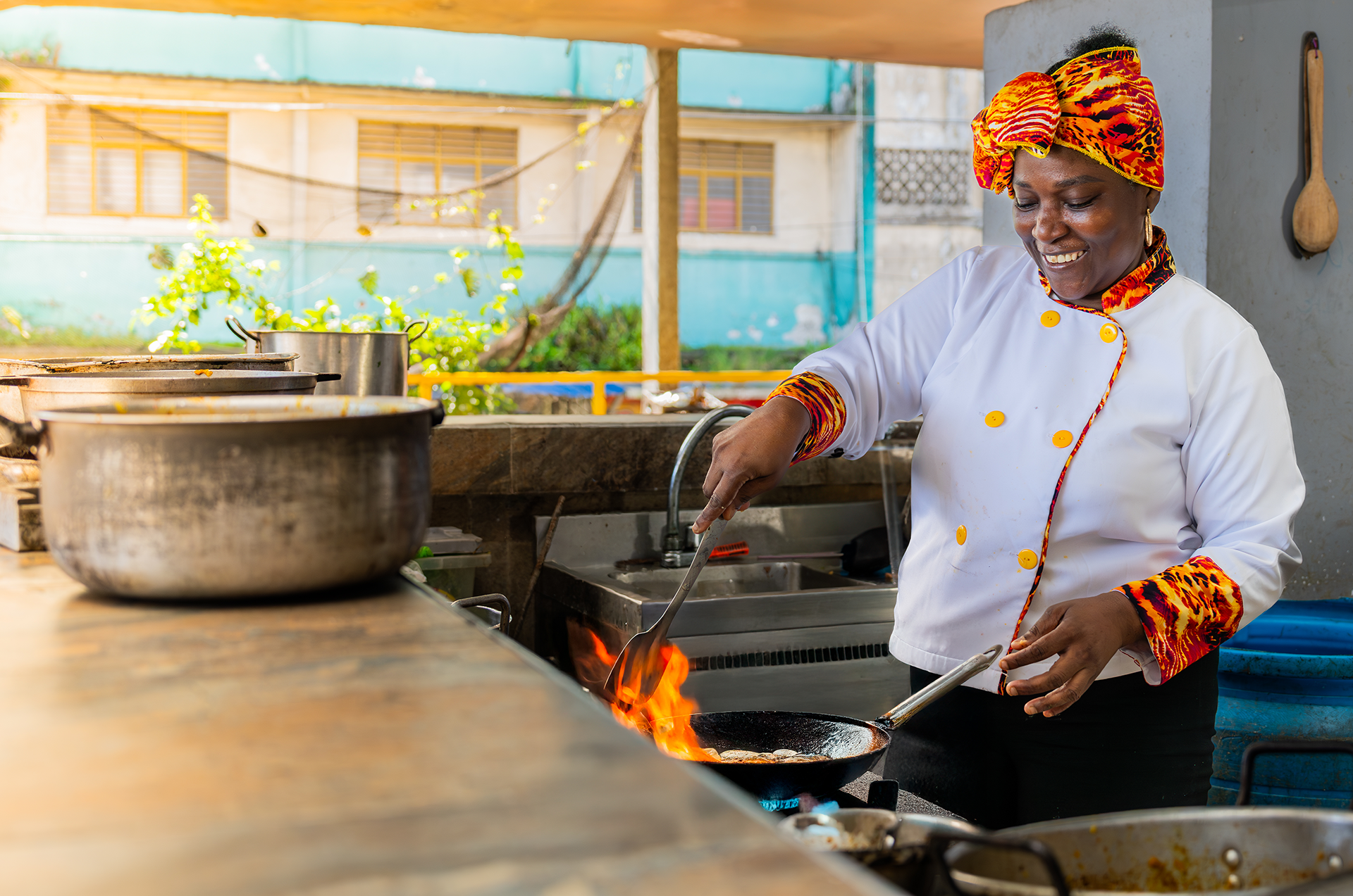 Mujer cocinando en una cocina al aire libre con ollas sobre el fuego. Ella viste una blusa blanca con detalles en naranja y un turbante colorido. Sonríe mientras cocina.