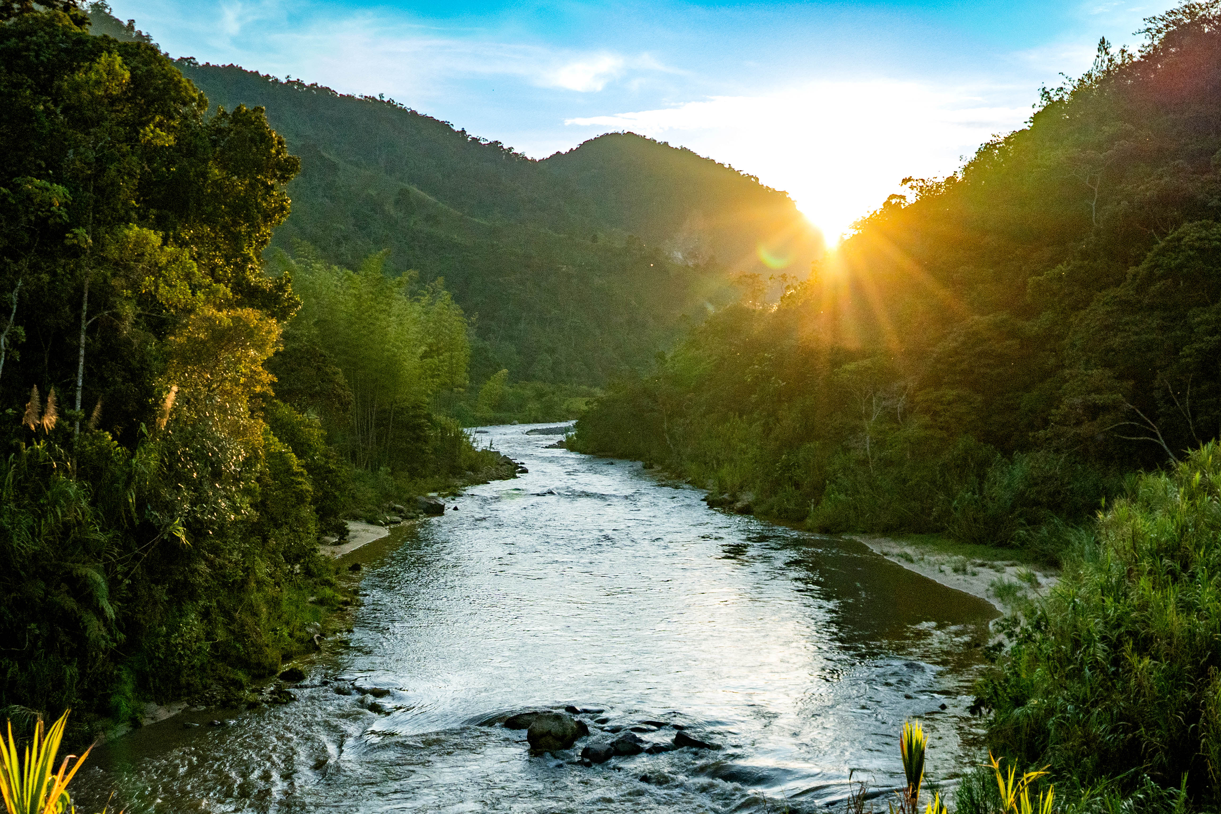 Un río que fluye entre un paisaje de montañas con vegetación densa y árboles verdes, mientras el sol se pone en el horizonte, creando un resplandor dorado en el cielo.