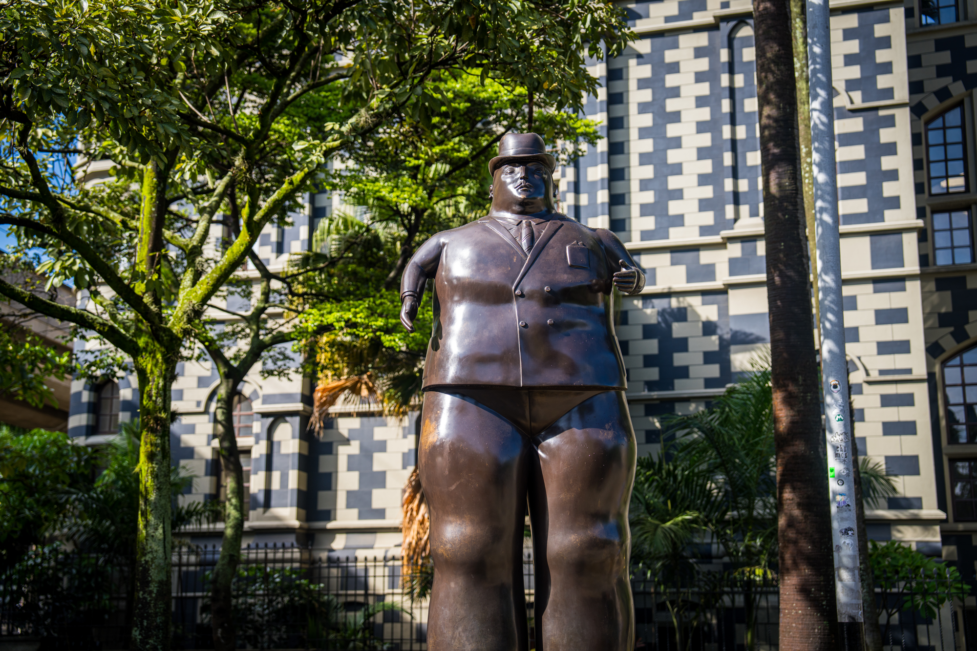 Estatua de un hombre con sombrero, traje y dejando una mano en el bolsillo, en un parque con árboles y un edificio moderno al fondo.