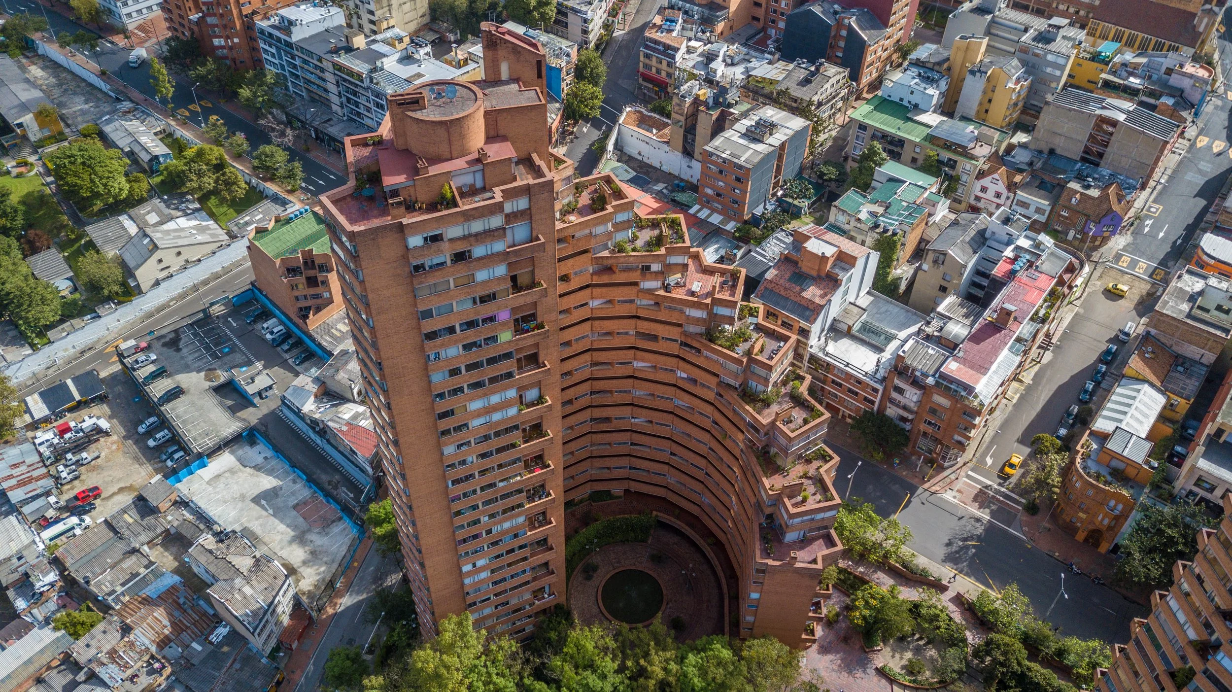 Vista aérea de una ciudad con edificios altos de ladrillo, calles con tráfico y áreas verdes, incluyendo un edificio prominente de estilo moderno con terrazas y plantas en la azotea.